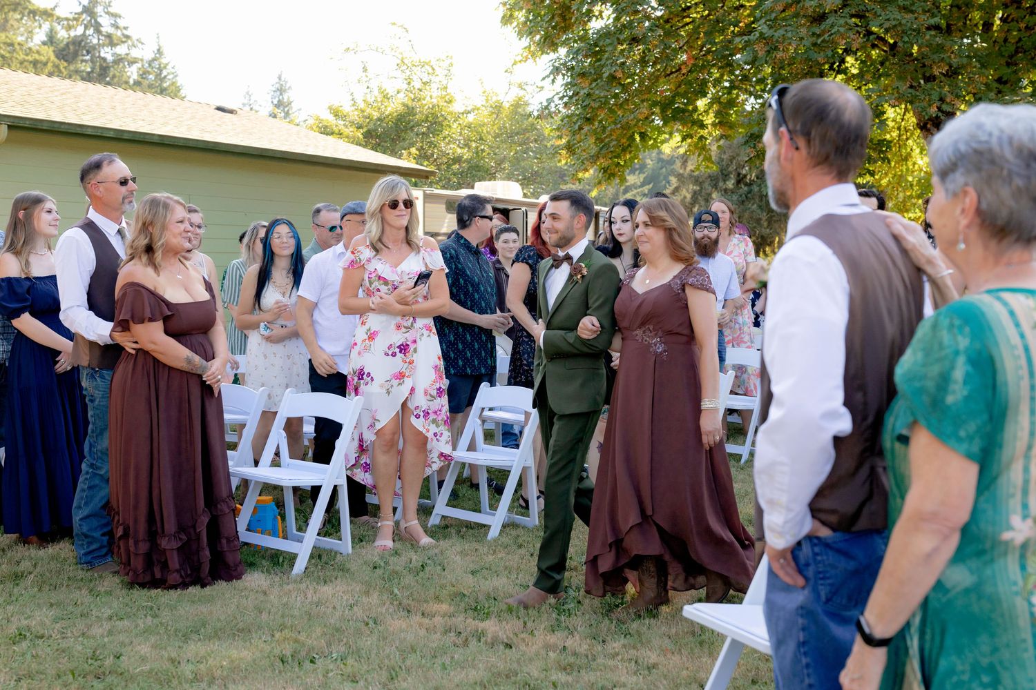 Wedding party in brown dresses and green suits gathered outdoors on sunny day.