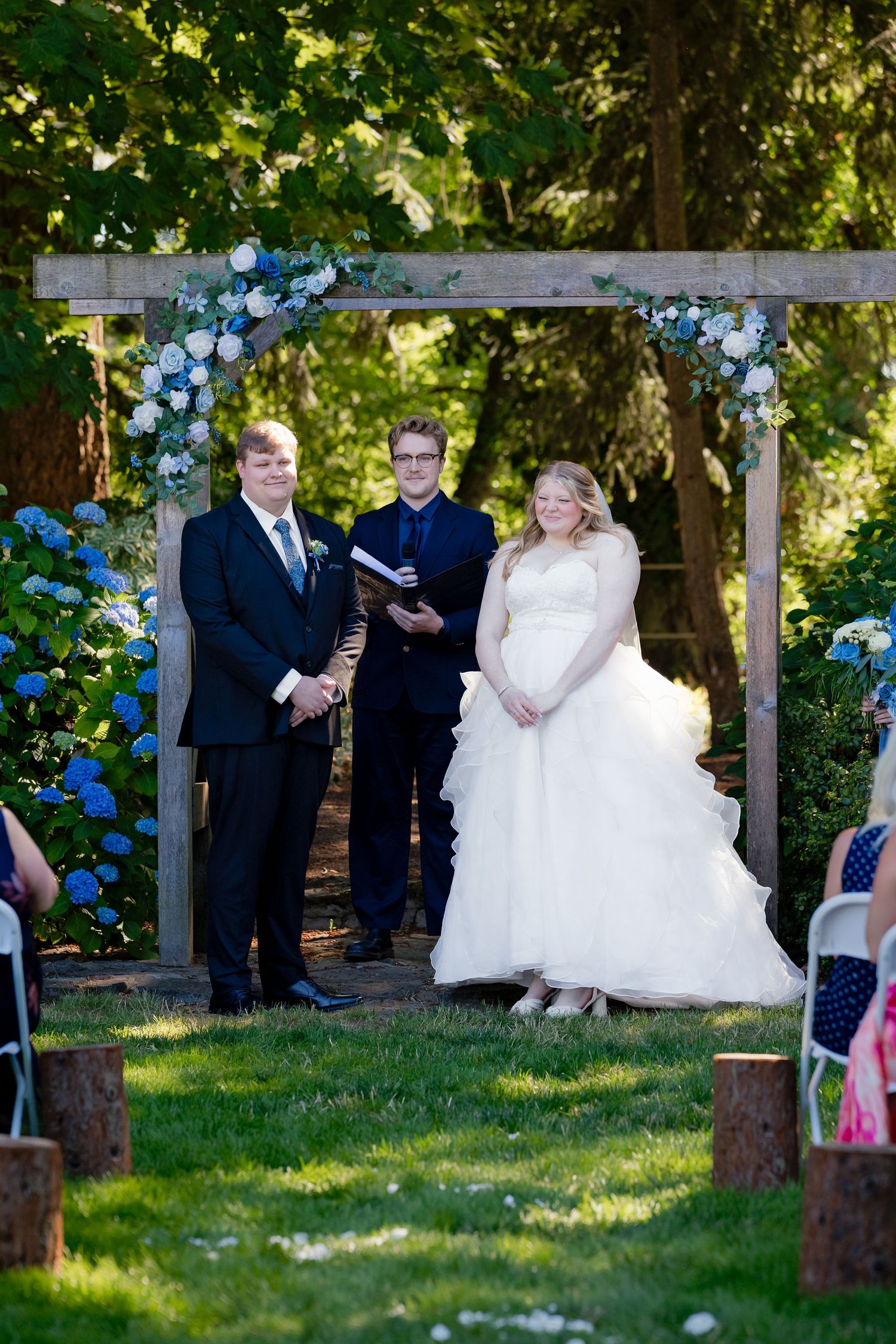 Bride and groom exchange vows under floral arch decorated with blue hydrangeas at outdoor garden wedding ceremony.