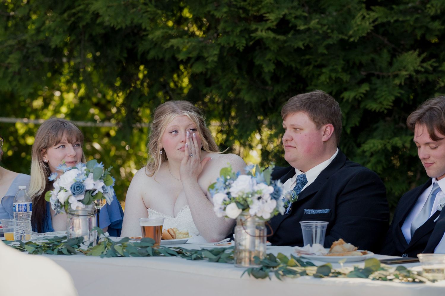 Bride wiping tears while seated at outdoor wedding reception head table decorated with white floral centerpieces.