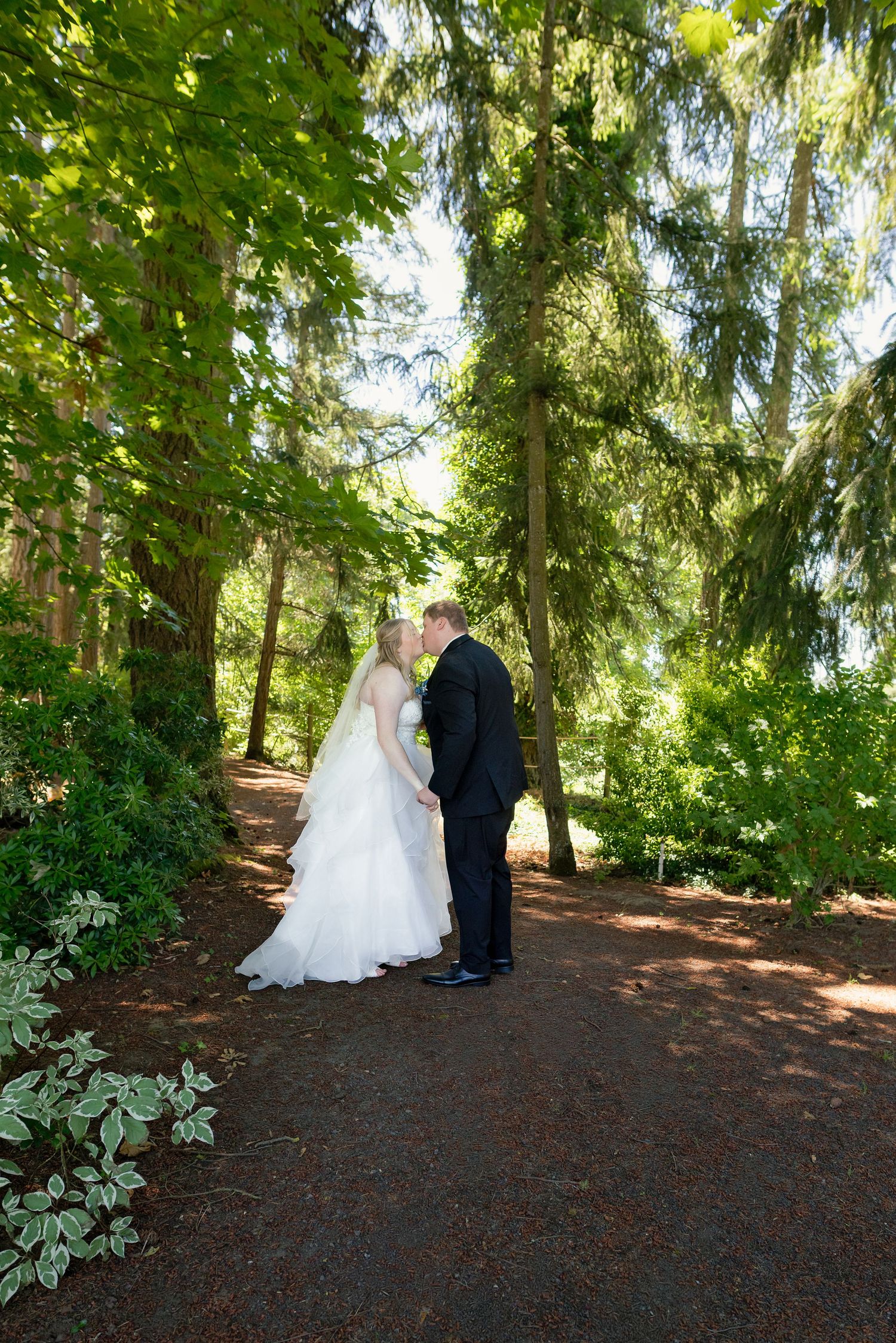 Bride and groom sharing a romantic moment on a tree-lined path surrounded by lush green foliage.