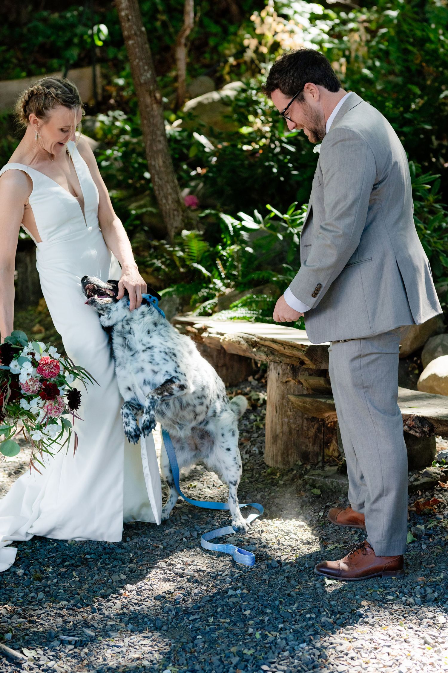 Bride and groom in gray suit interact with their spotted dog on a gravel path during wedding photos.