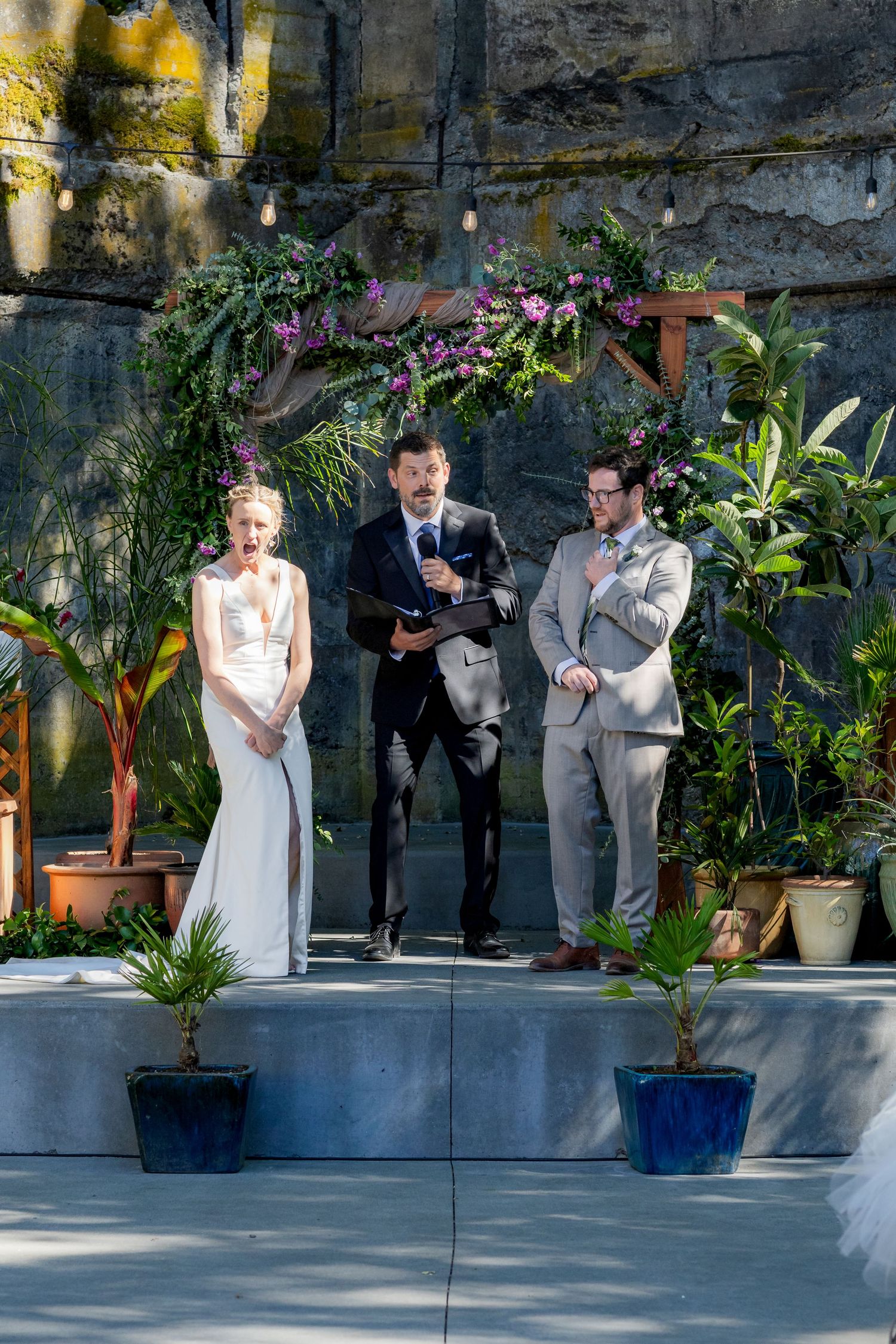 Bride and groom stand at outdoor wedding altar with floral arch as officiant reads vows.