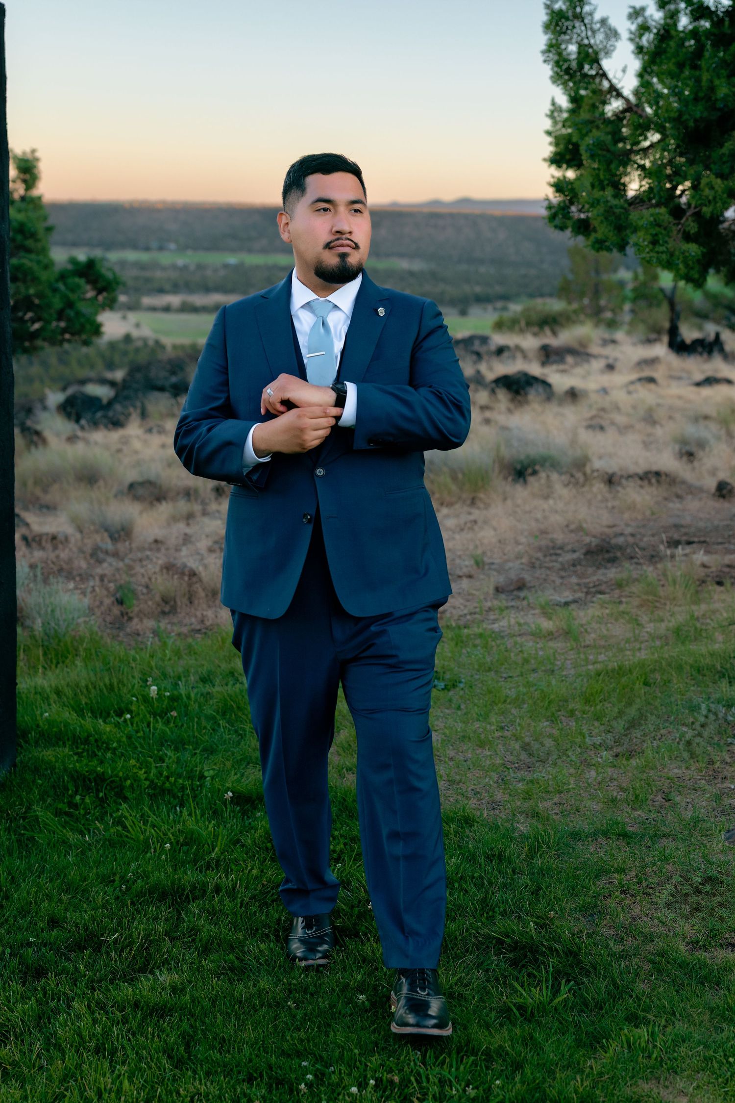 Groom in navy blue suit adjusts cufflinks at sunset with scenic landscape behind.