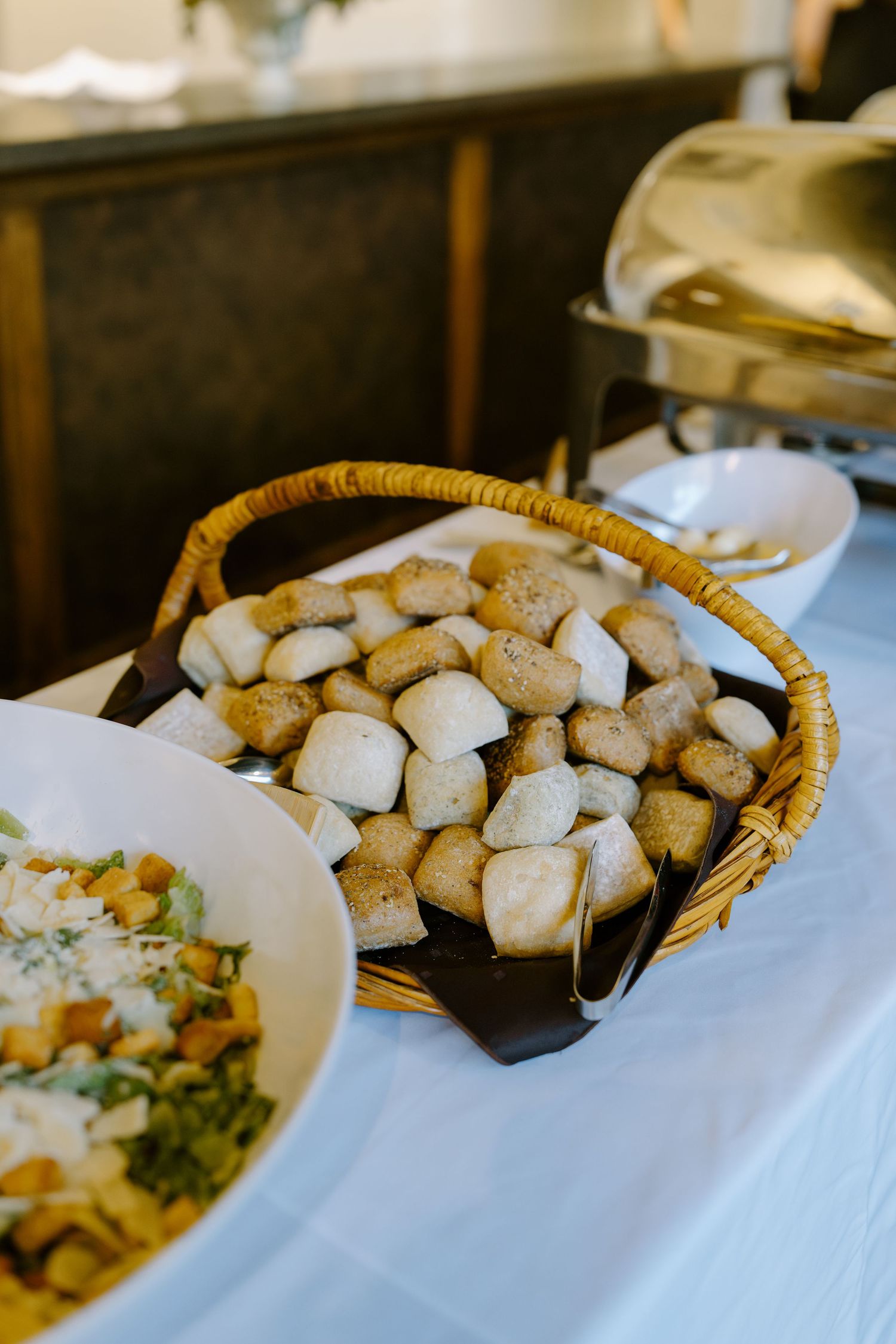 Wicker basket filled with dinner rolls on white tablecloth beside Caesar salad at wedding reception buffet.