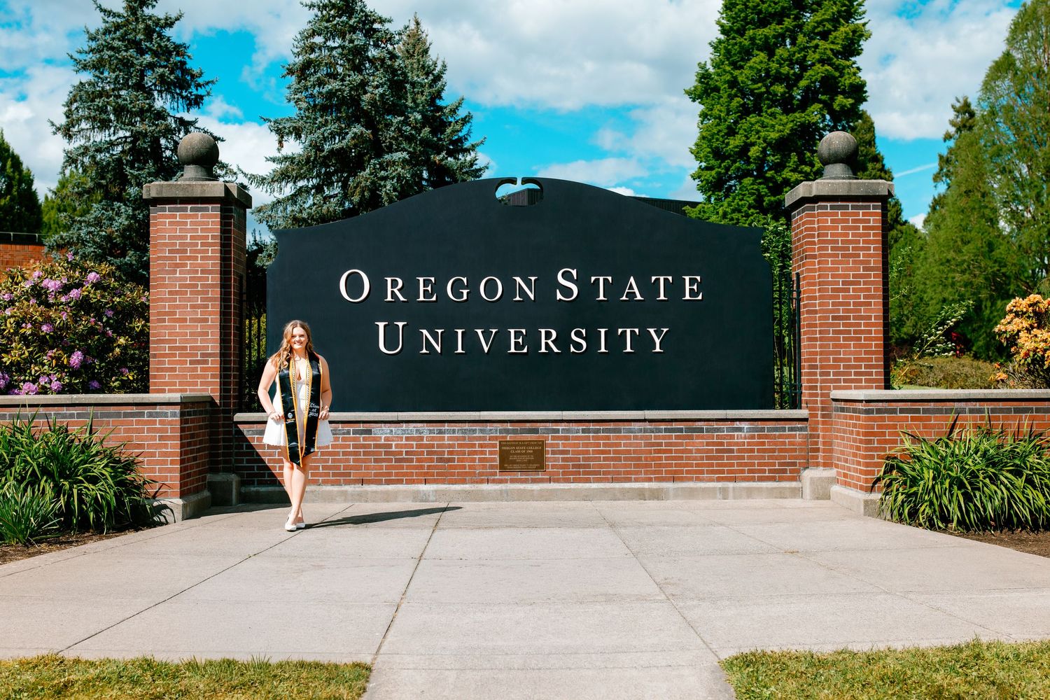 Graduate poses in cap and gown by the Oregon State University brick entrance sign on a sunny day.