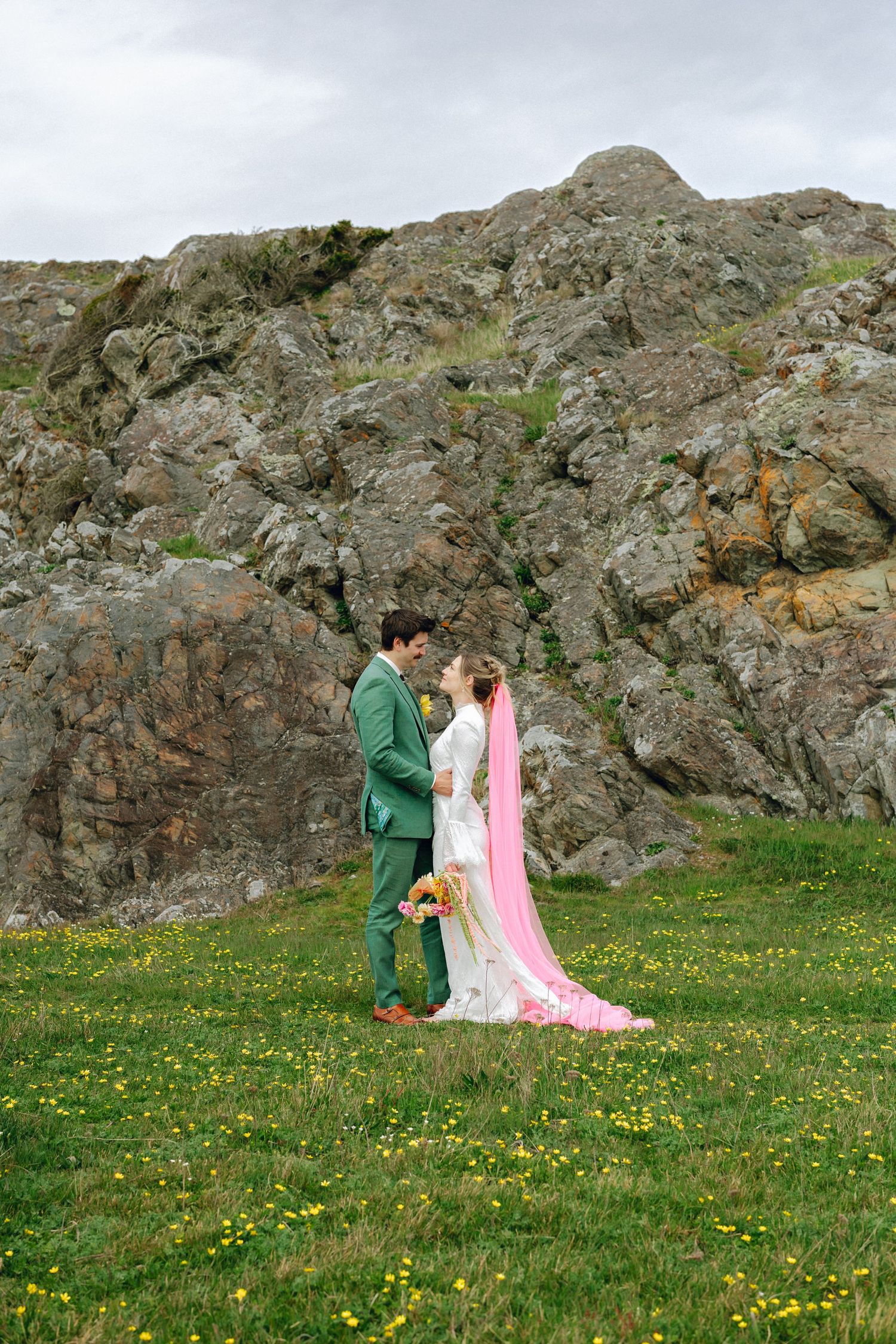 Bride in white gown with pink veil and groom in green suit embrace in wildflower meadow before rocky cliffs.
