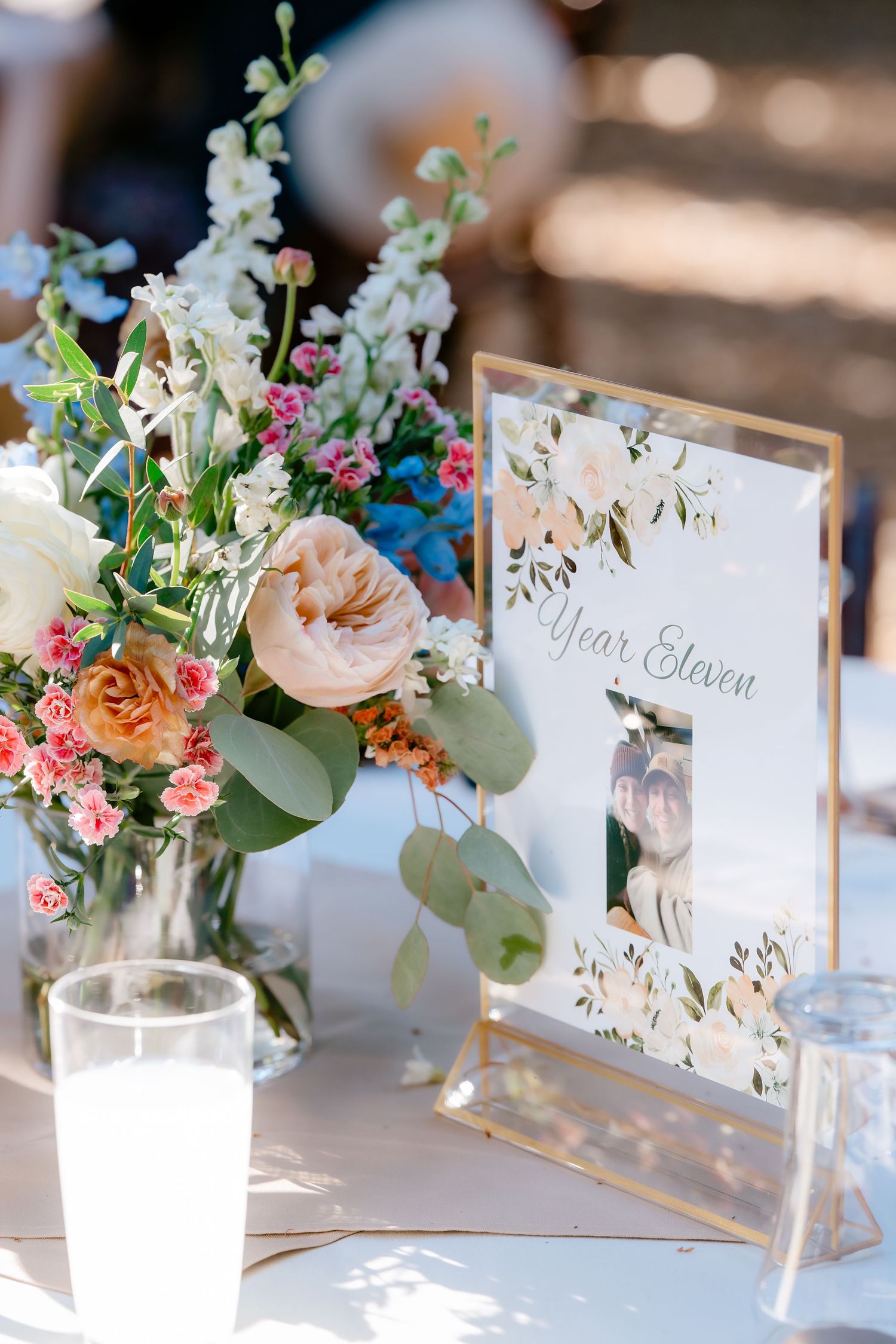 Elegant wedding table centerpiece with pink and white floral arrangement beside a Year Eleven table sign in a gold frame.
