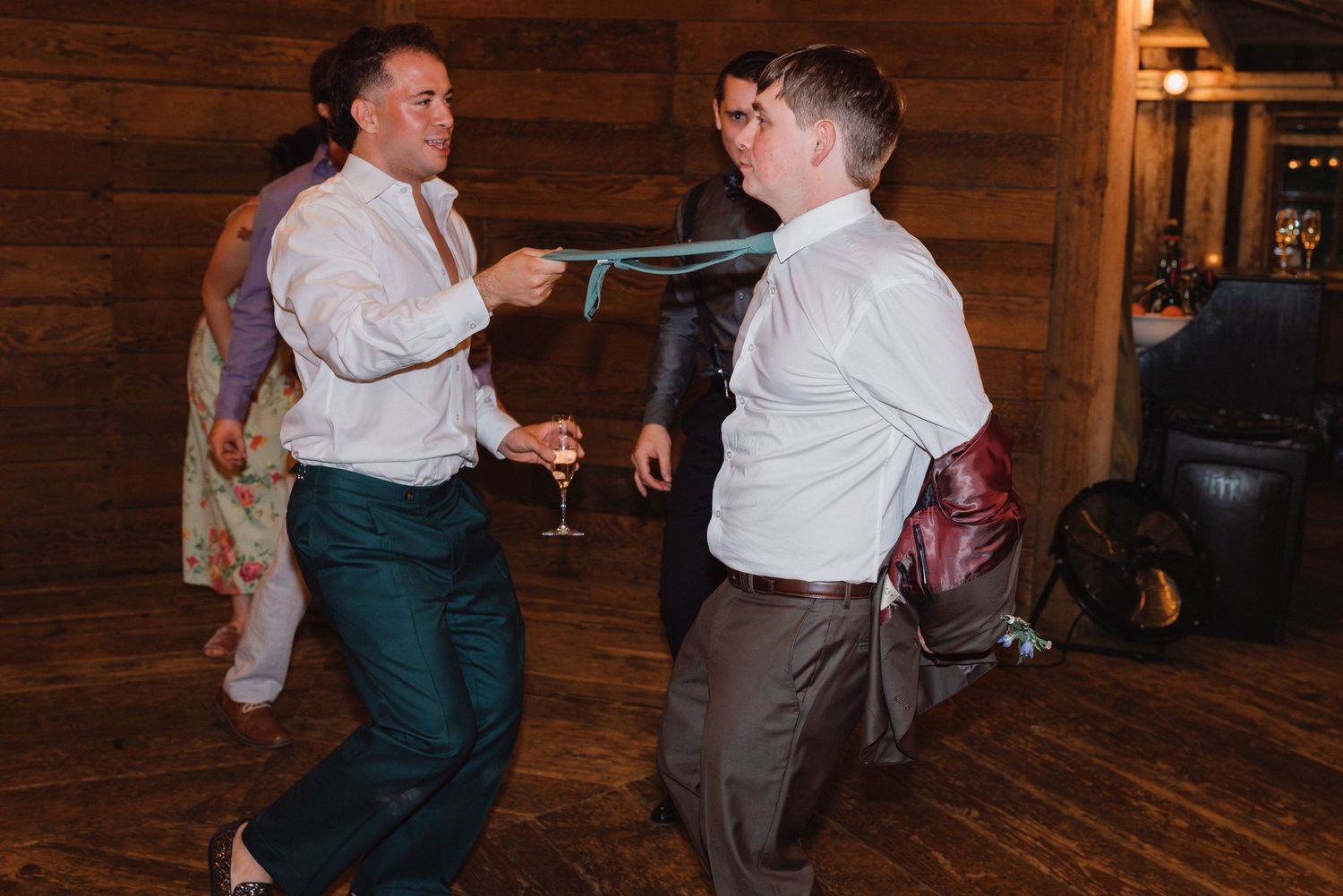 Two men playfully dancing and pulling a tie during a lively indoor wedding reception party.