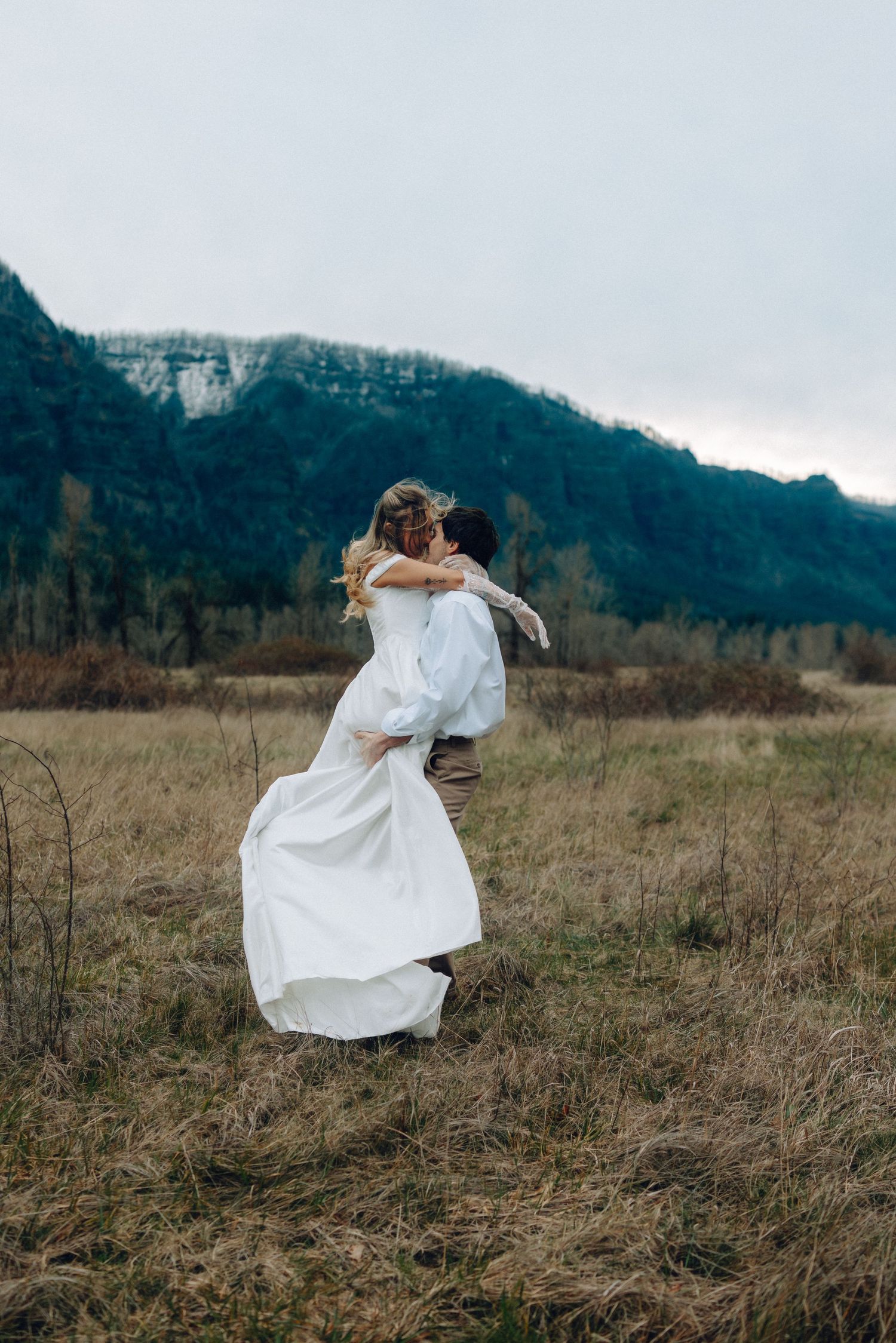 Couple in wedding attire dancing in a rustic field with mountains in the background.