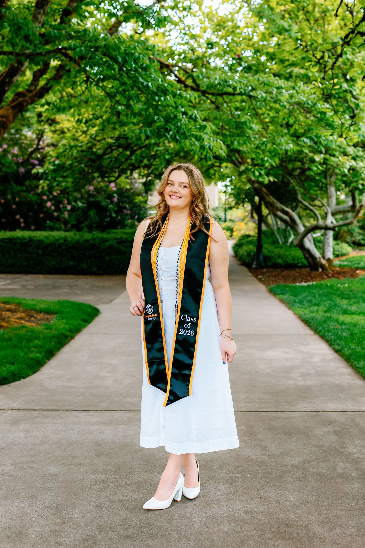 A smiling graduate in a white dress and green Class of 2020 sash poses on a tree-lined campus walkway.