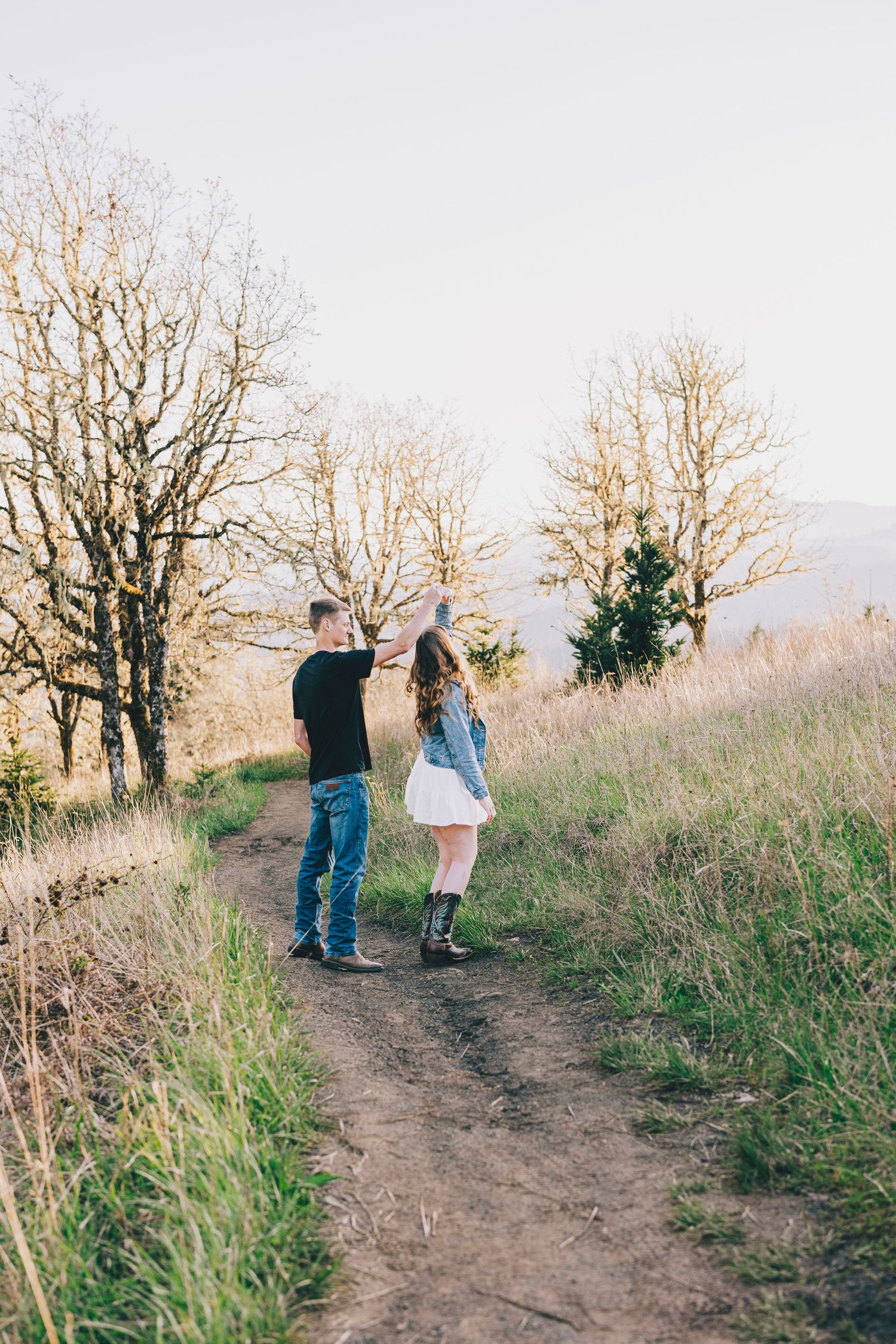 Two people walk hand in hand along a dirt trail surrounded by bare trees and autumn grass.