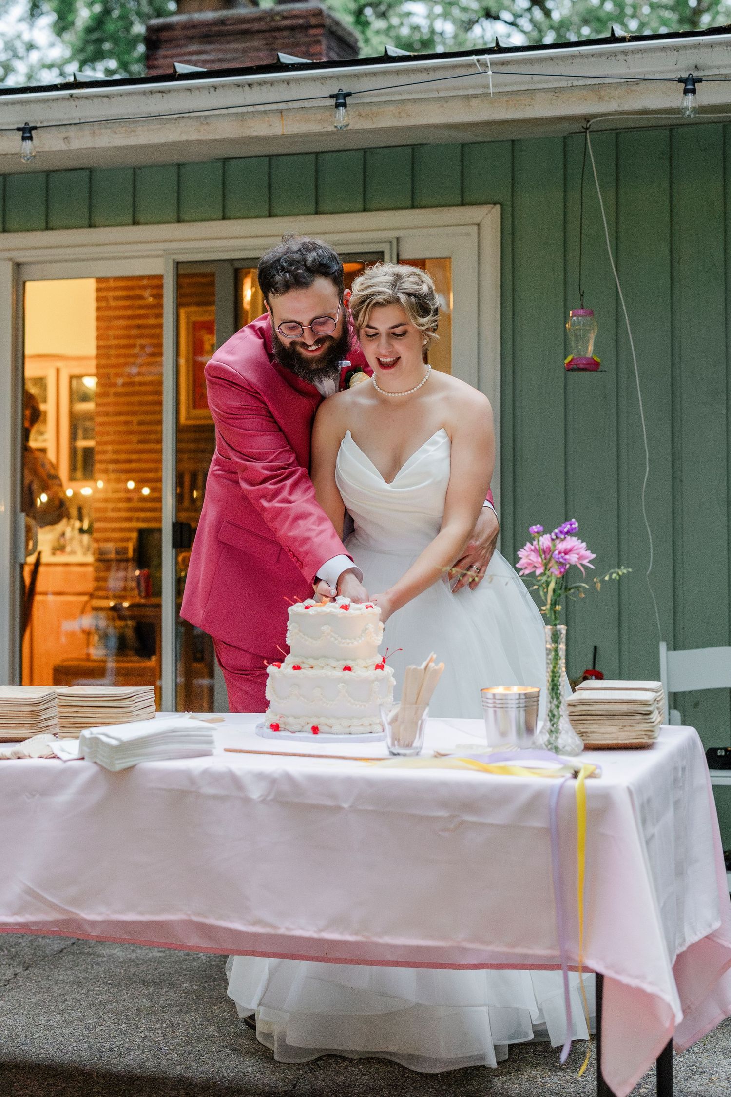 Couple cuts their wedding cake together at an outdoor reception with a white tablecloth and green backdrop.