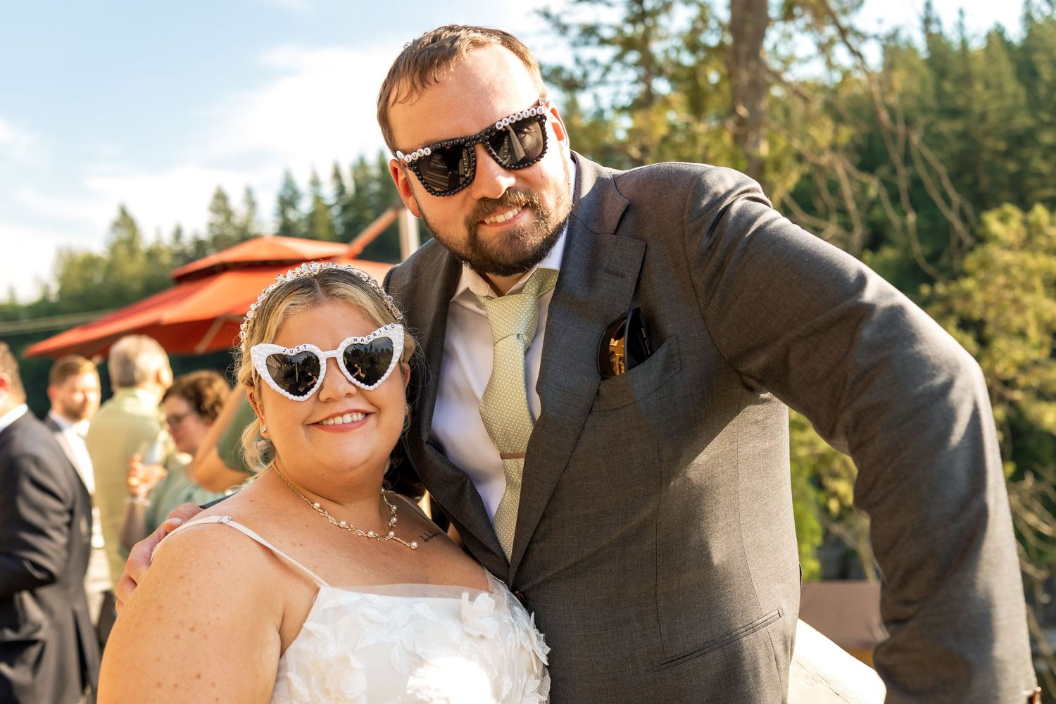 Couple wearing sunglasses poses for casual outdoor portrait with forest backdrop.