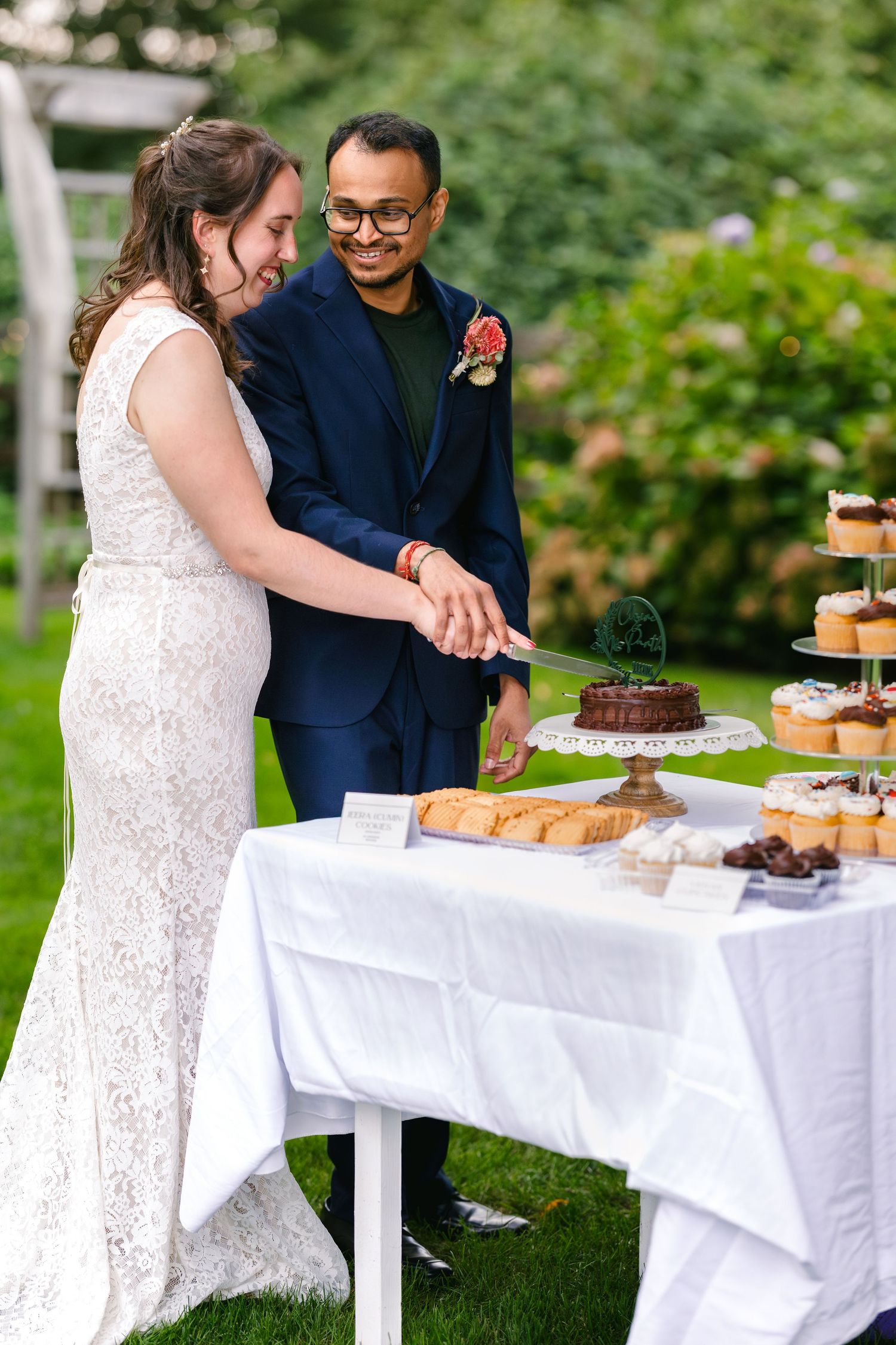 Newlyweds share a moment while cutting their wedding cake at an outdoor dessert table.