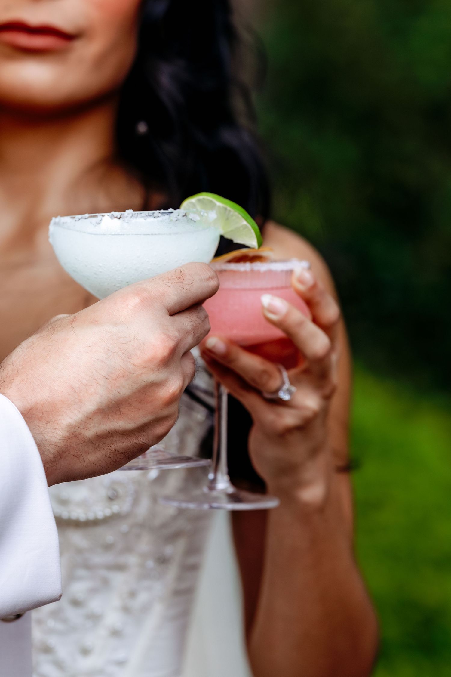 Close-up of cocktail glasses with lime garnish being held at an outdoor wedding celebration.
