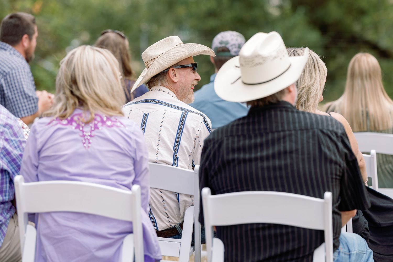 Wedding guests in cowboy hats and casual attire seated in white chairs at an outdoor ceremony.