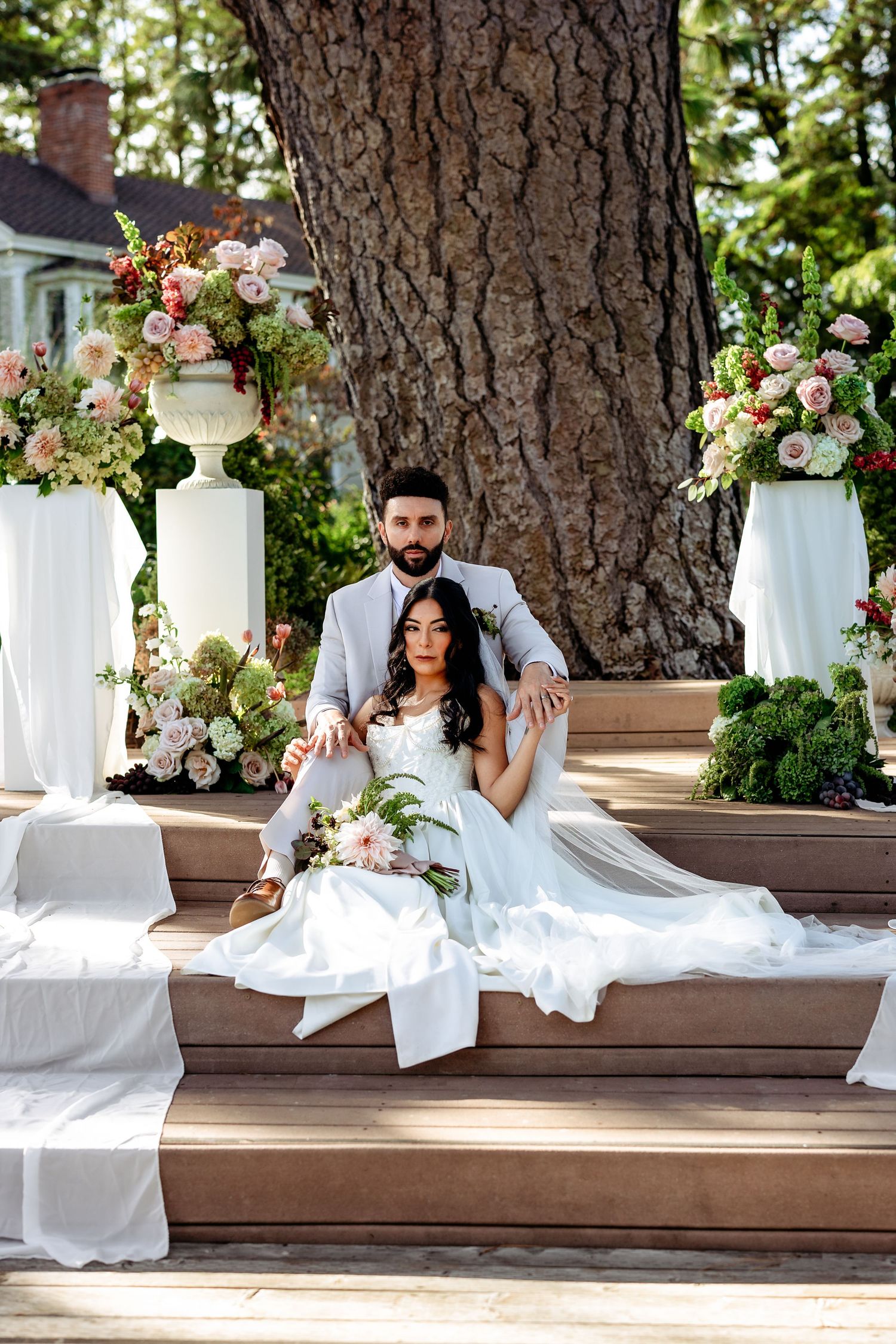 A romantic wedding couple poses on wooden steps surrounded by lush greenery and floral arrangements.