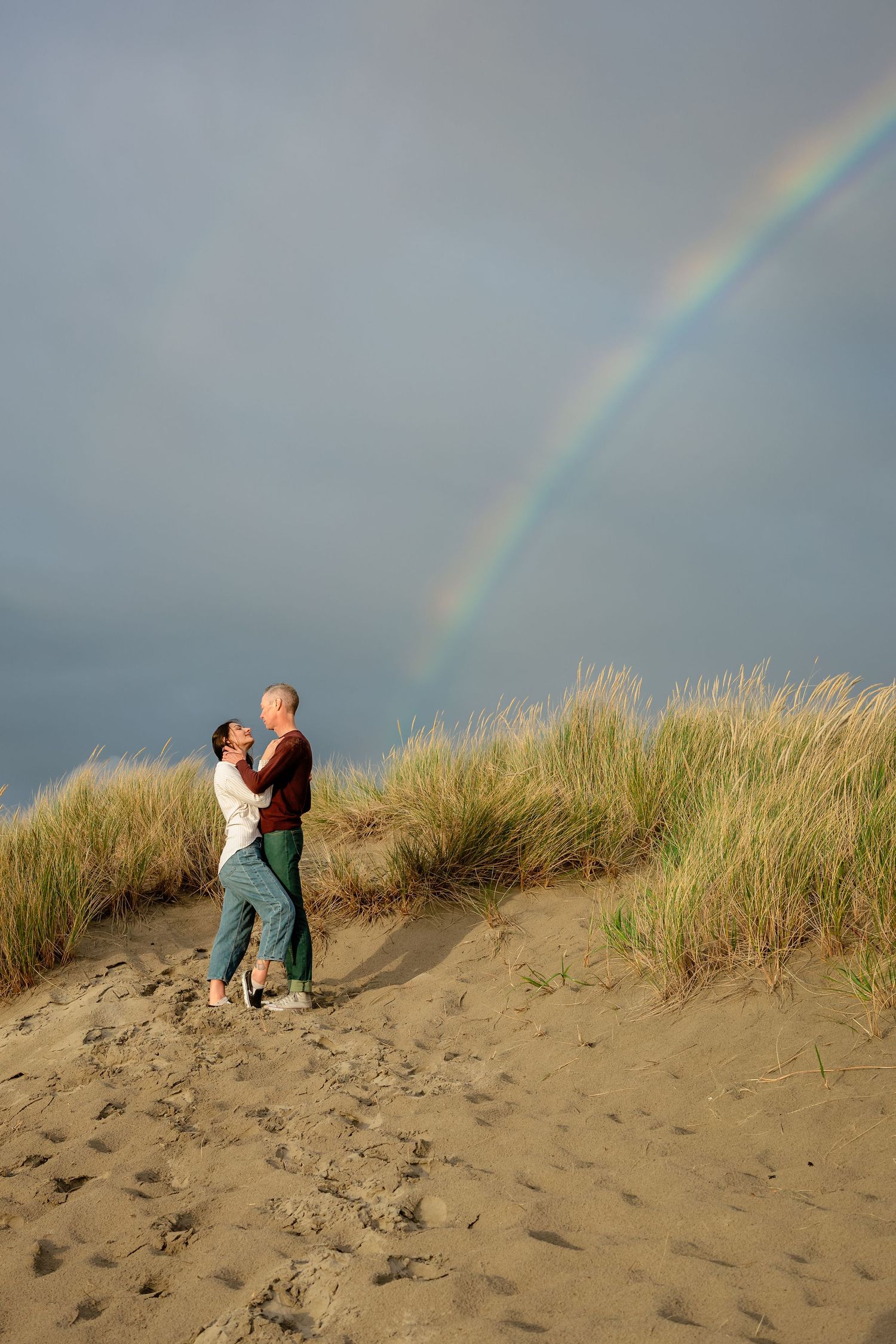 A rainbow appears over sandy beach dunes with tall grass as people explore the scenic coastal landscape.