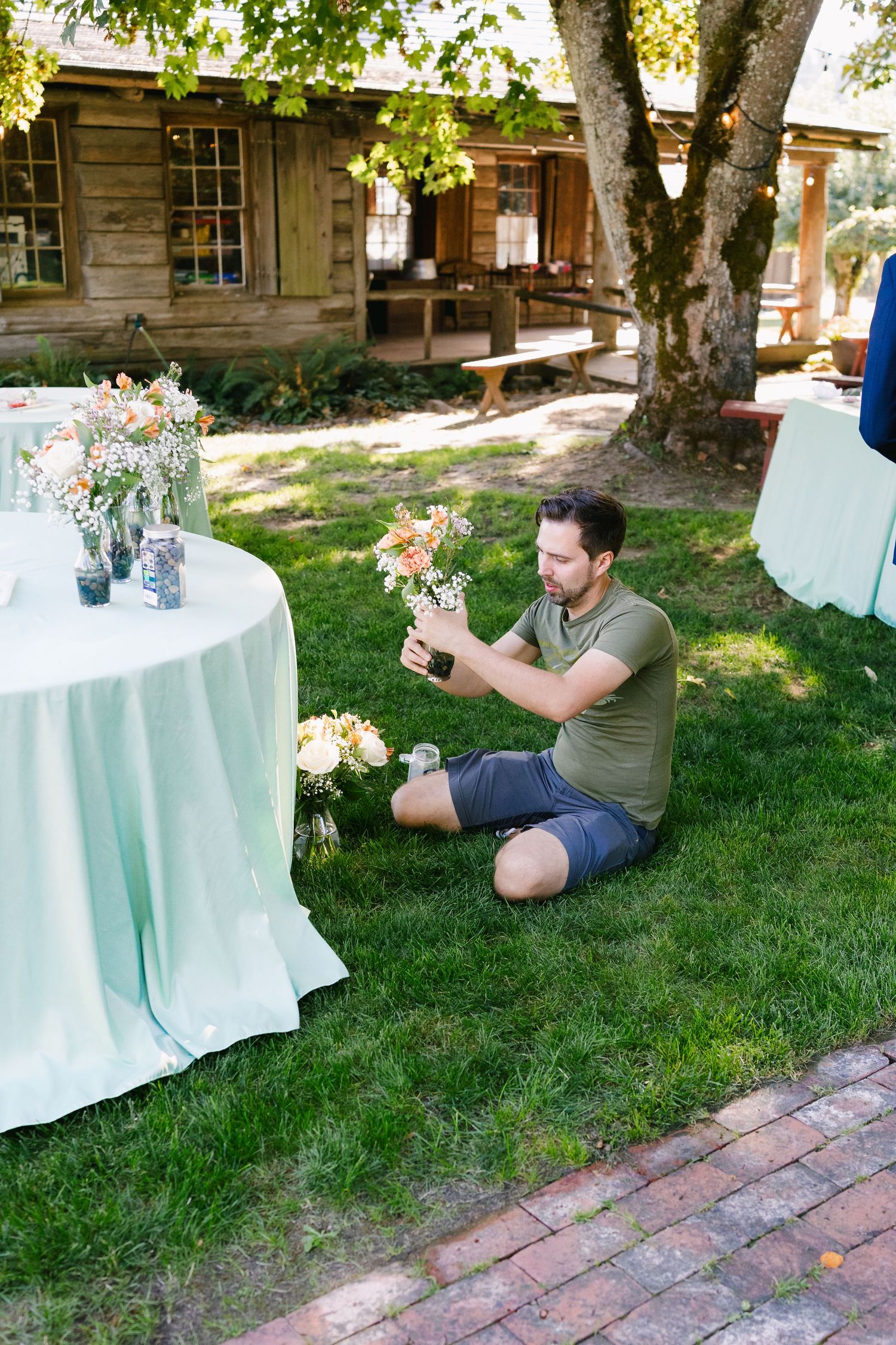 A garden scene with a round table covered in mint green cloth and floral arrangements near a rustic cabin.
