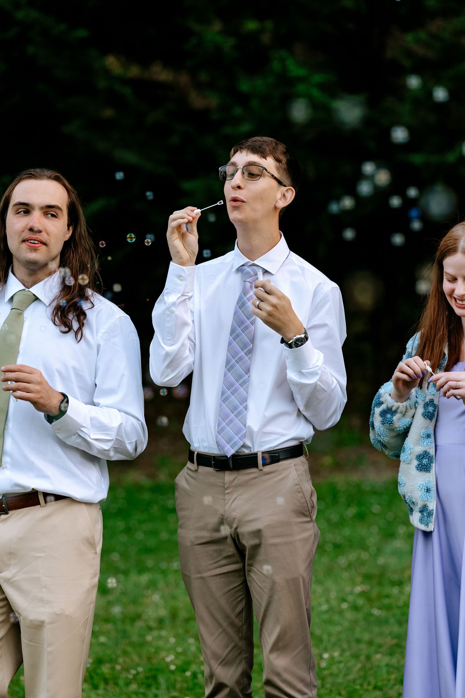 Wedding guests blow bubbles outdoors during a celebration on a lush green lawn at dusk.