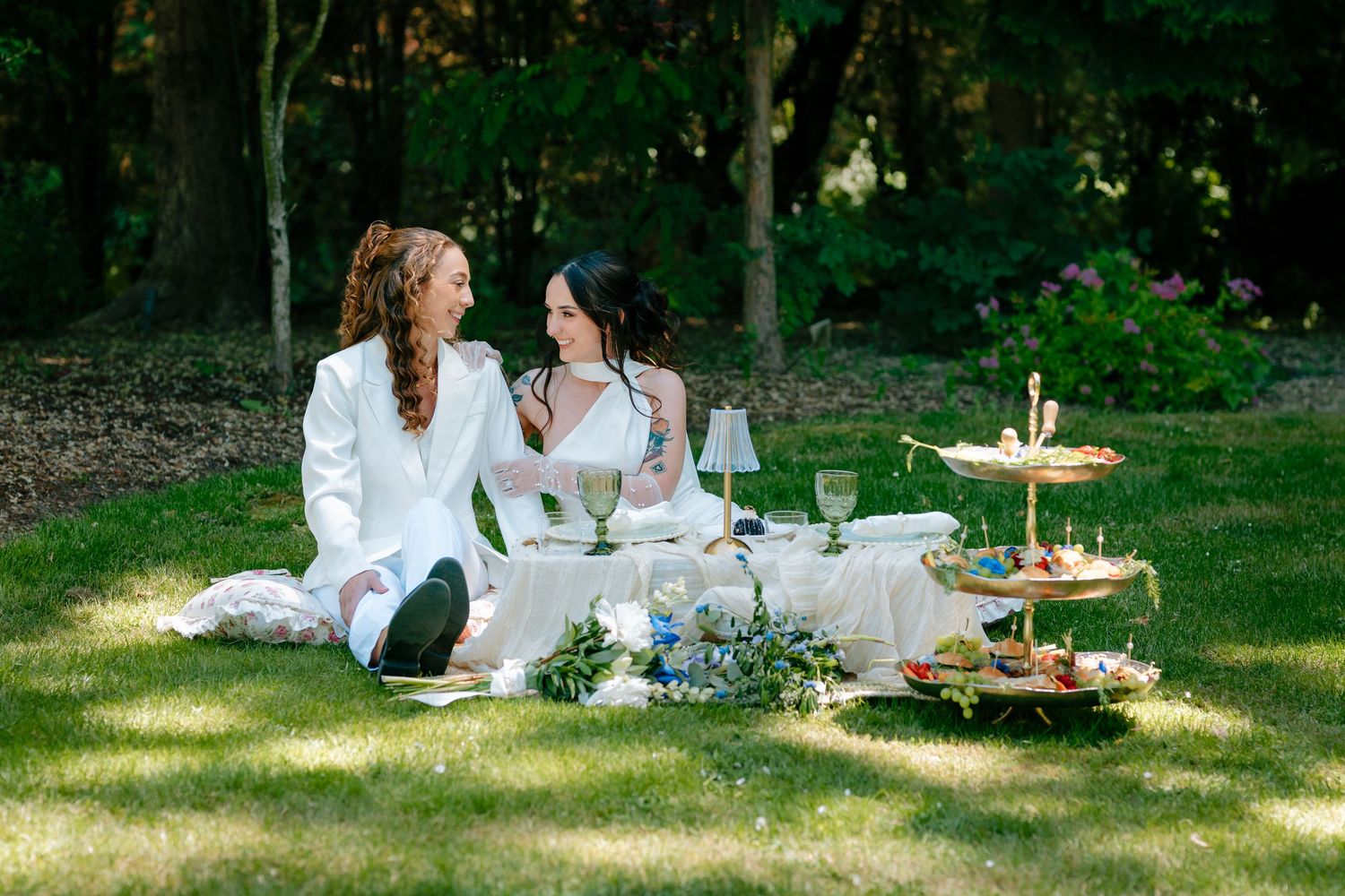 Two brides in white sit on grass enjoying an elegant outdoor picnic with tiered dessert stand.