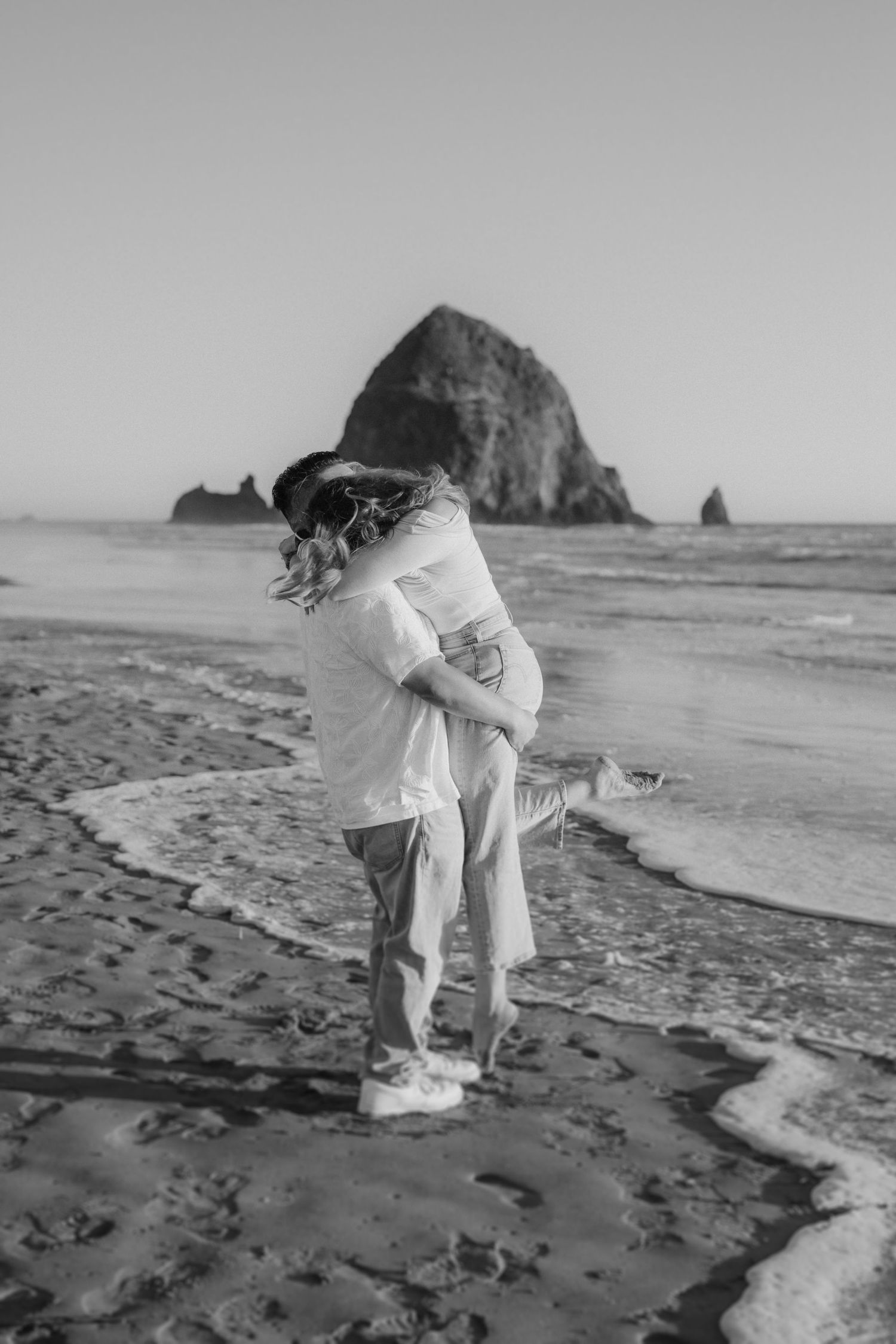 Black and white photo of a couple embracing on a beach near Haystack Rock at Cannon Beach, Oregon.