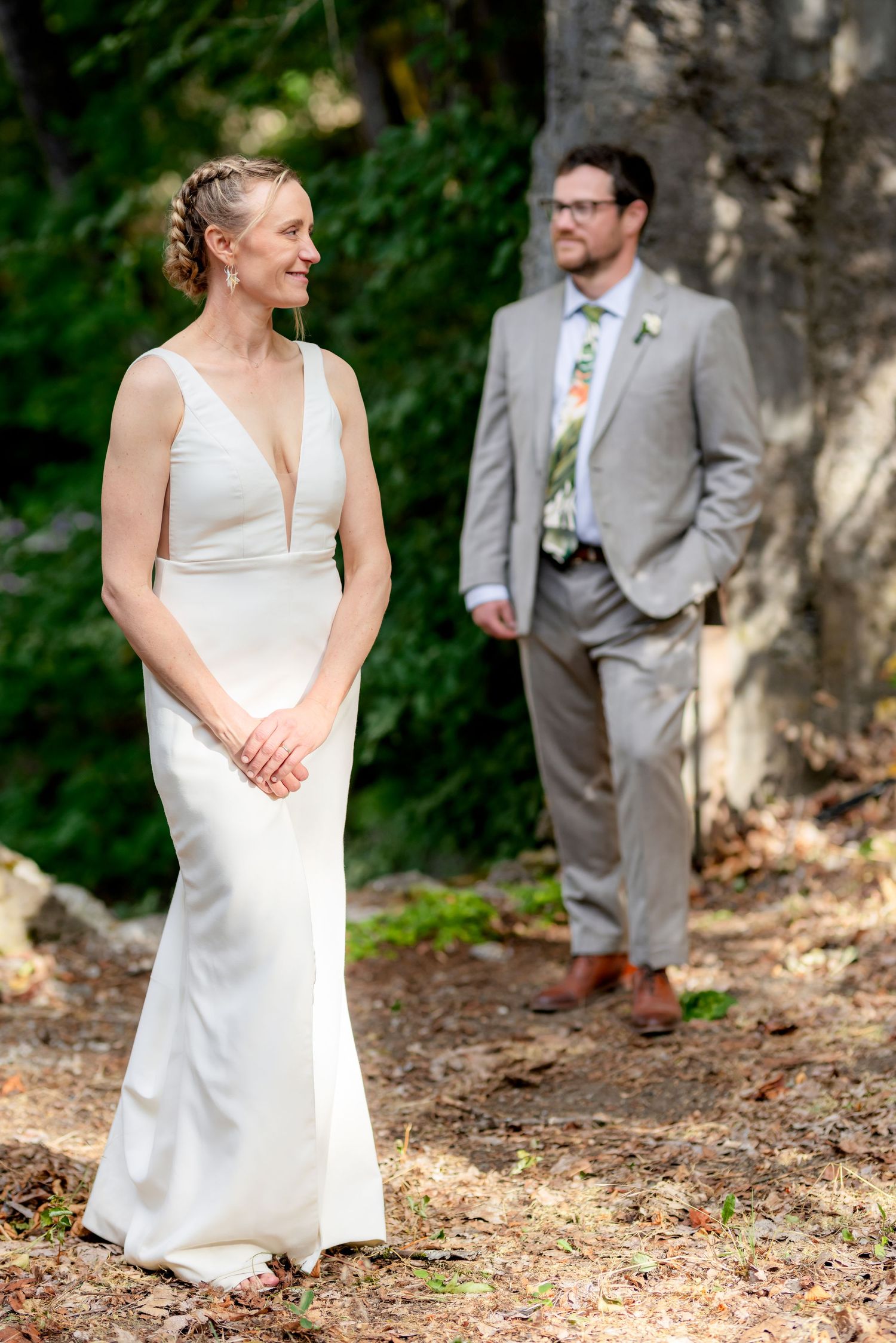 Bride in white gown smiles outdoors as groom in gray suit stands behind her among trees at wedding.