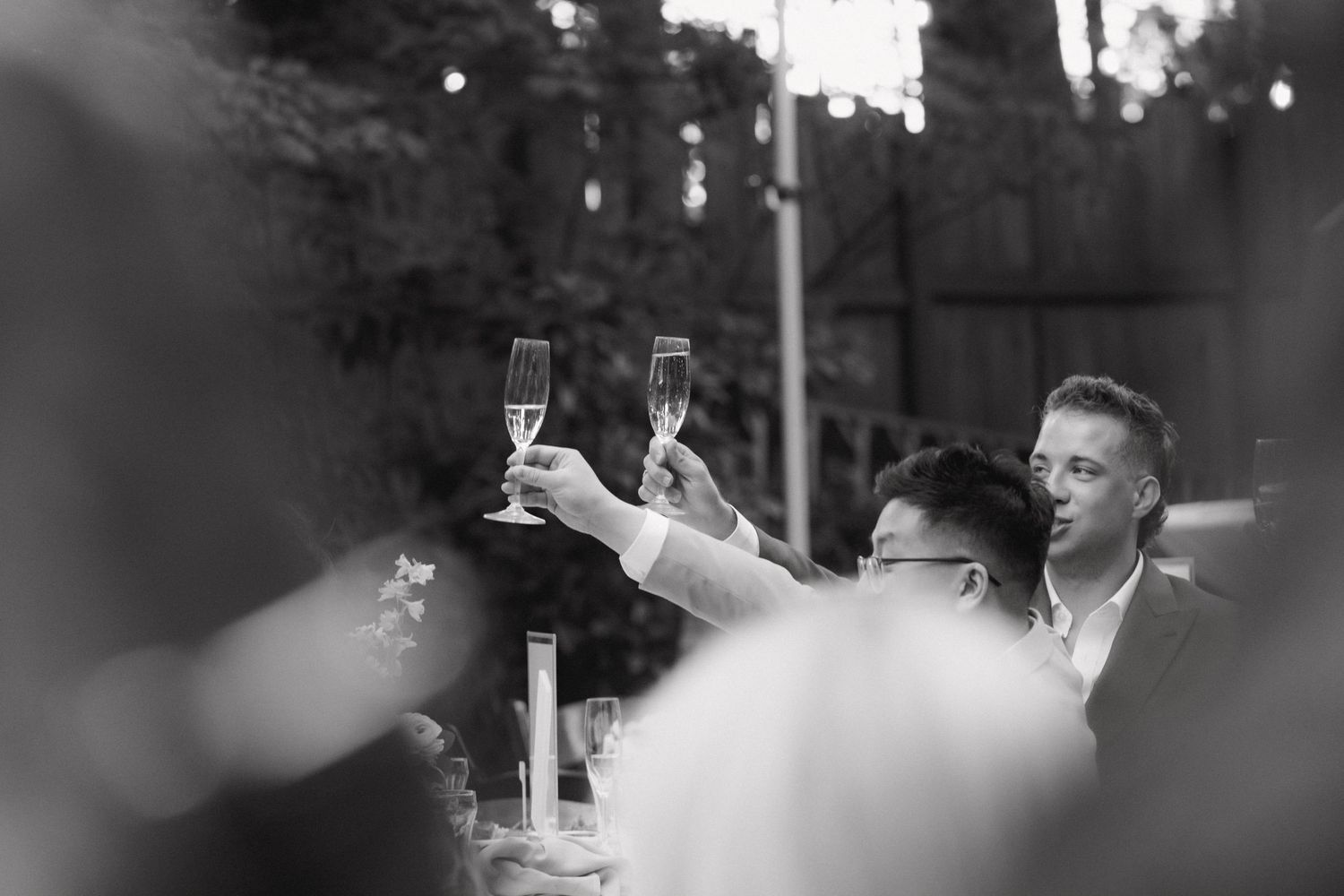 Black and white photo of two men raising champagne glasses in toast at wedding reception.