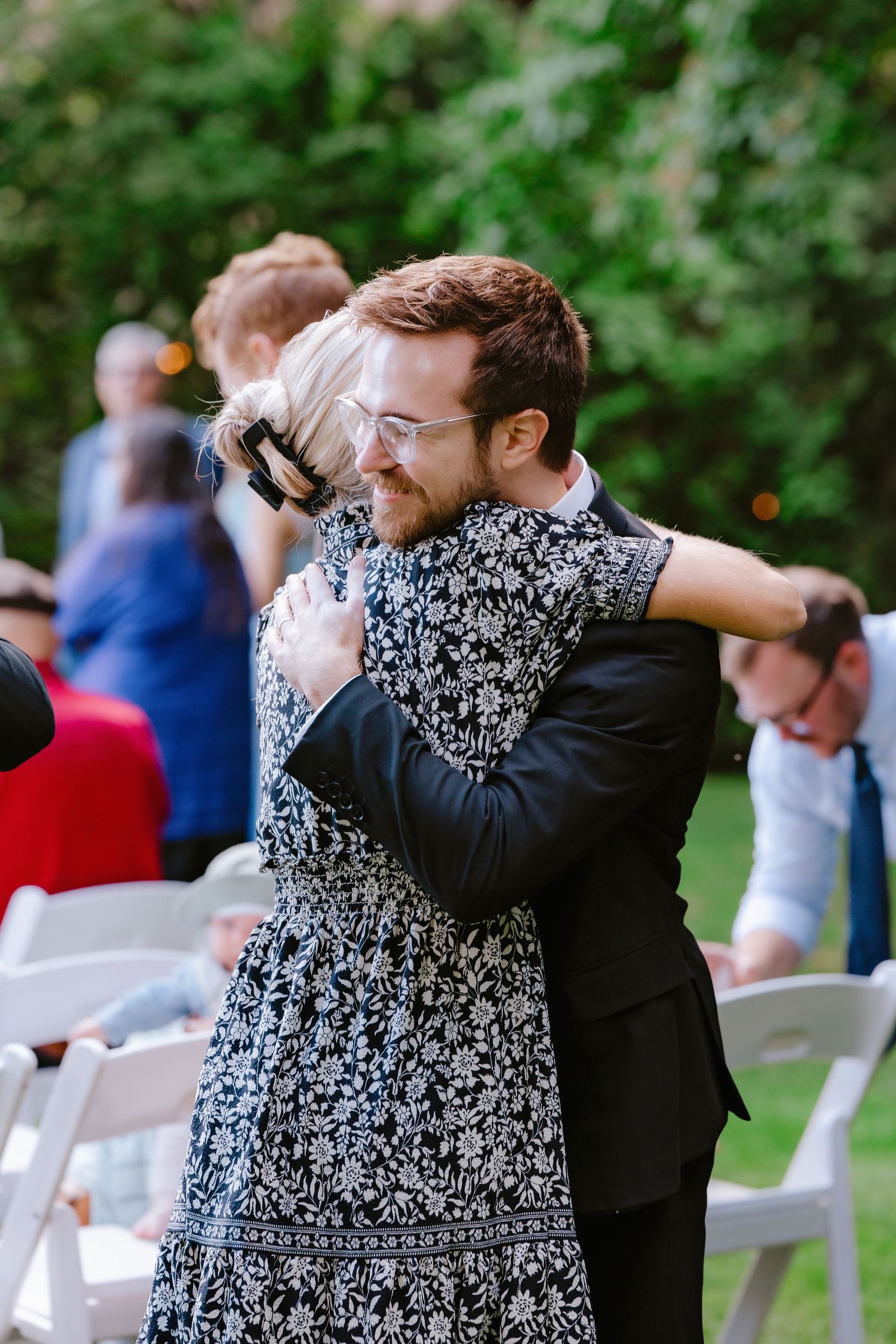 A man with glasses and beard hugs a woman in a floral dress at an outdoor wedding ceremony.