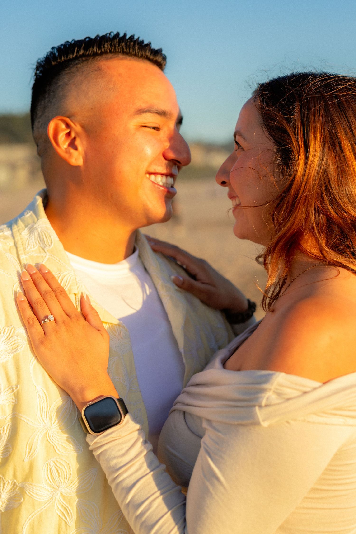 Smiling couple gazing at each other during golden hour beach sunset, woman wearing engagement ring.