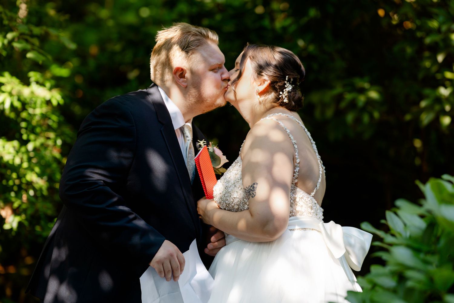 Bride and groom share a romantic first kiss outdoors surrounded by lush green trees on their wedding day.