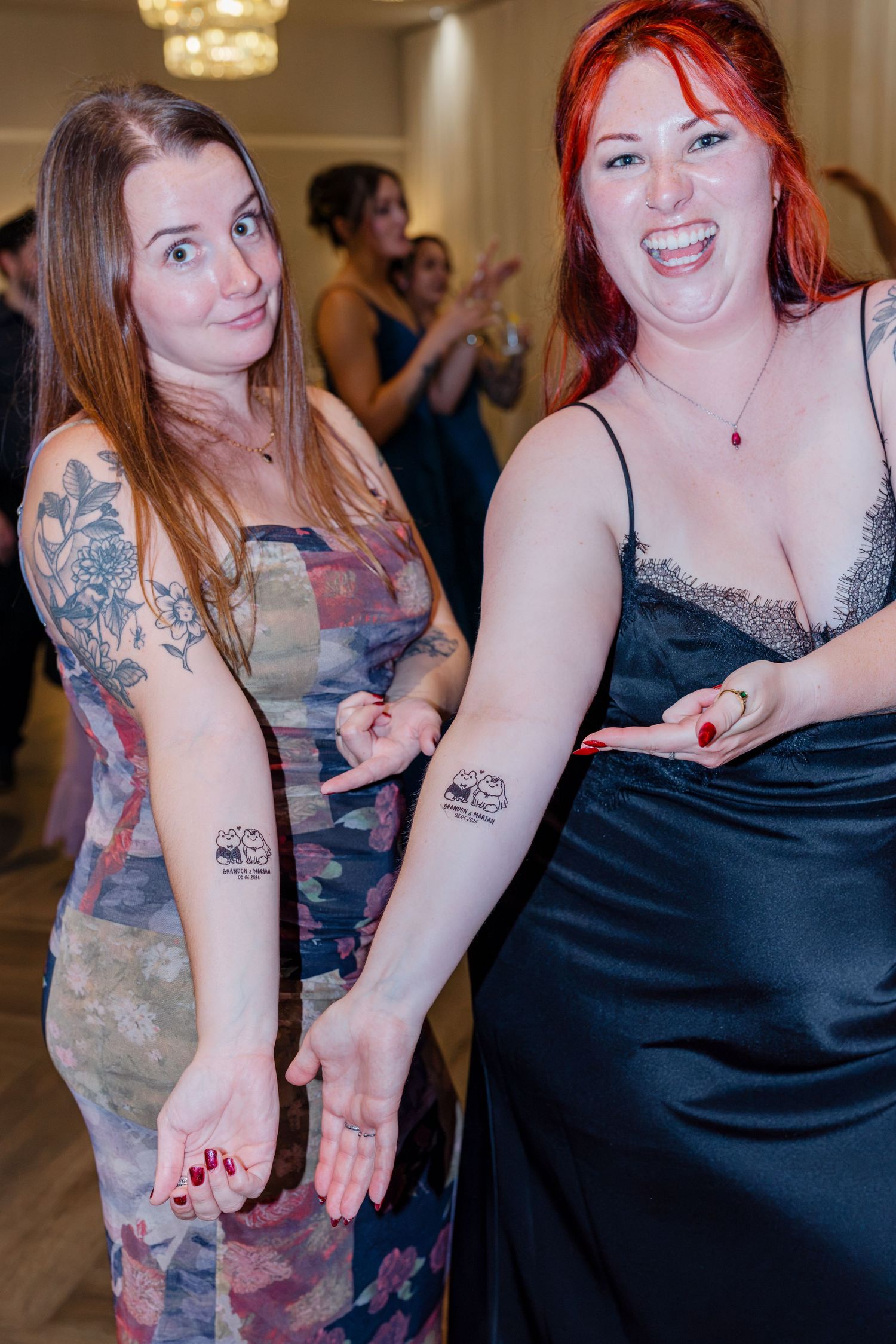 Two smiling women show off matching temporary tattoos on their arms at a wedding reception celebration.