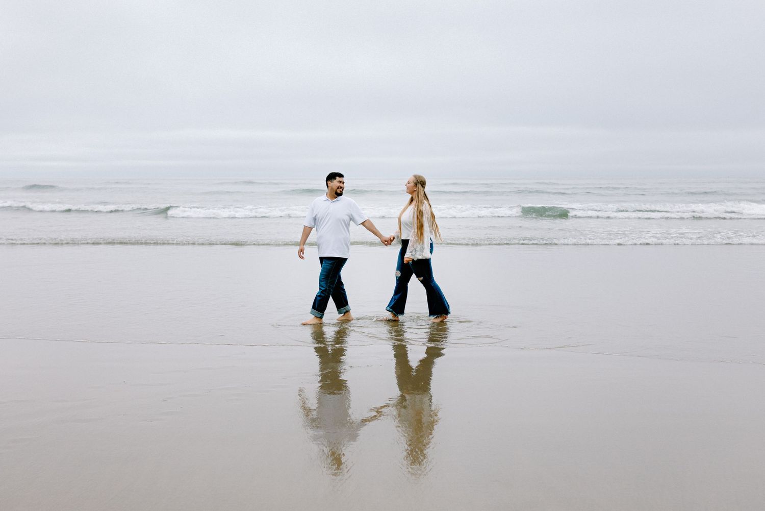 Couple holding hands and walking on a reflective wet beach with ocean waves and overcast sky.