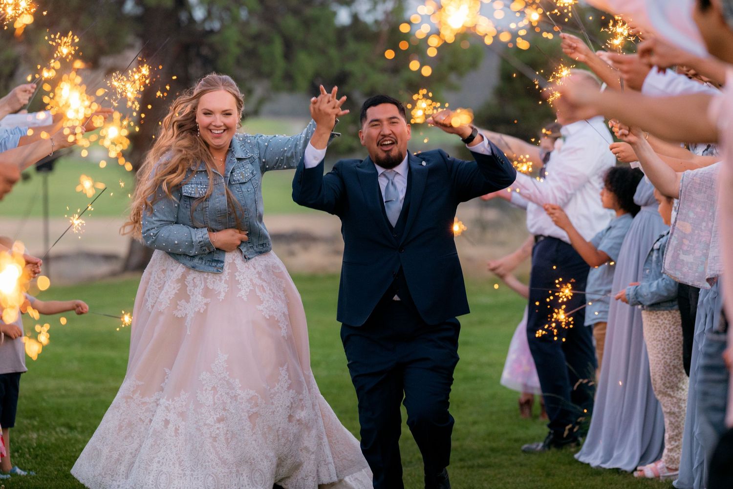 Newlyweds celebrate with sparkler exit as bride in white gown and denim jacket walks with groom.