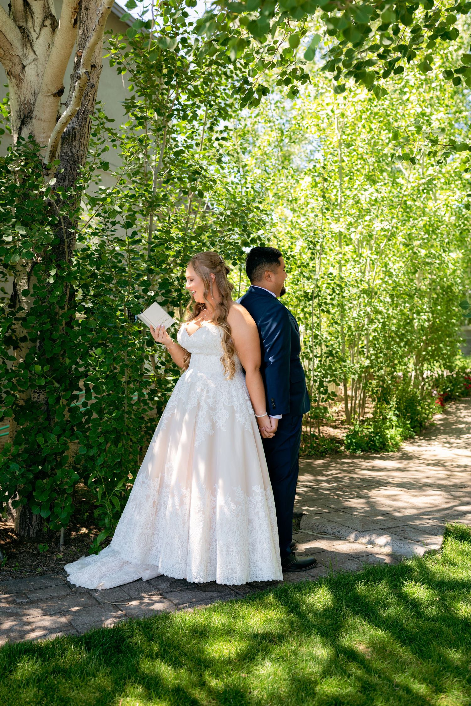 Bride in lace ball gown reads vows while groom stands back-to-back in blue suit during outdoor first look.