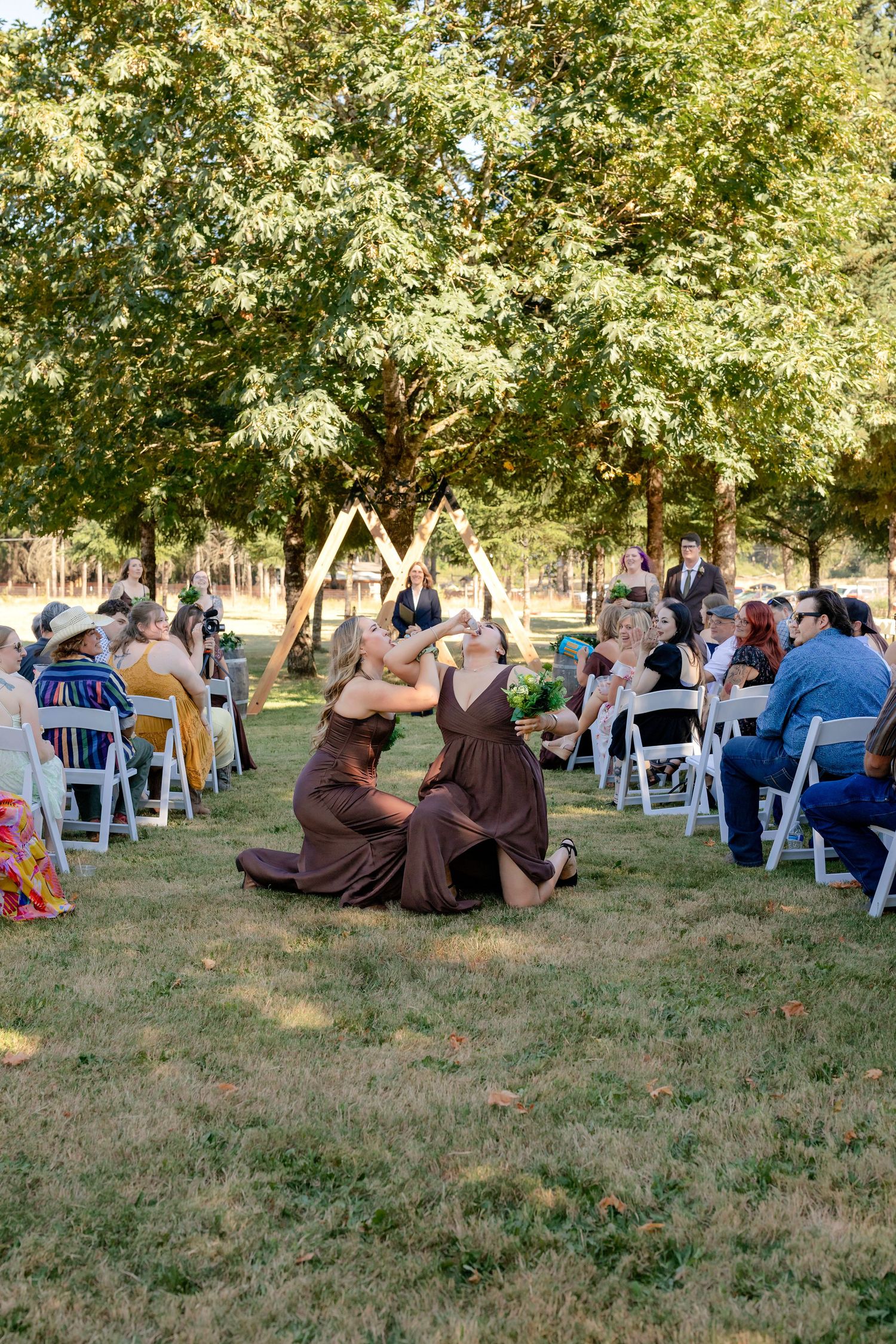 Two bridesmaids in brown dresses sitting on grass during an outdoor wedding ceremony.