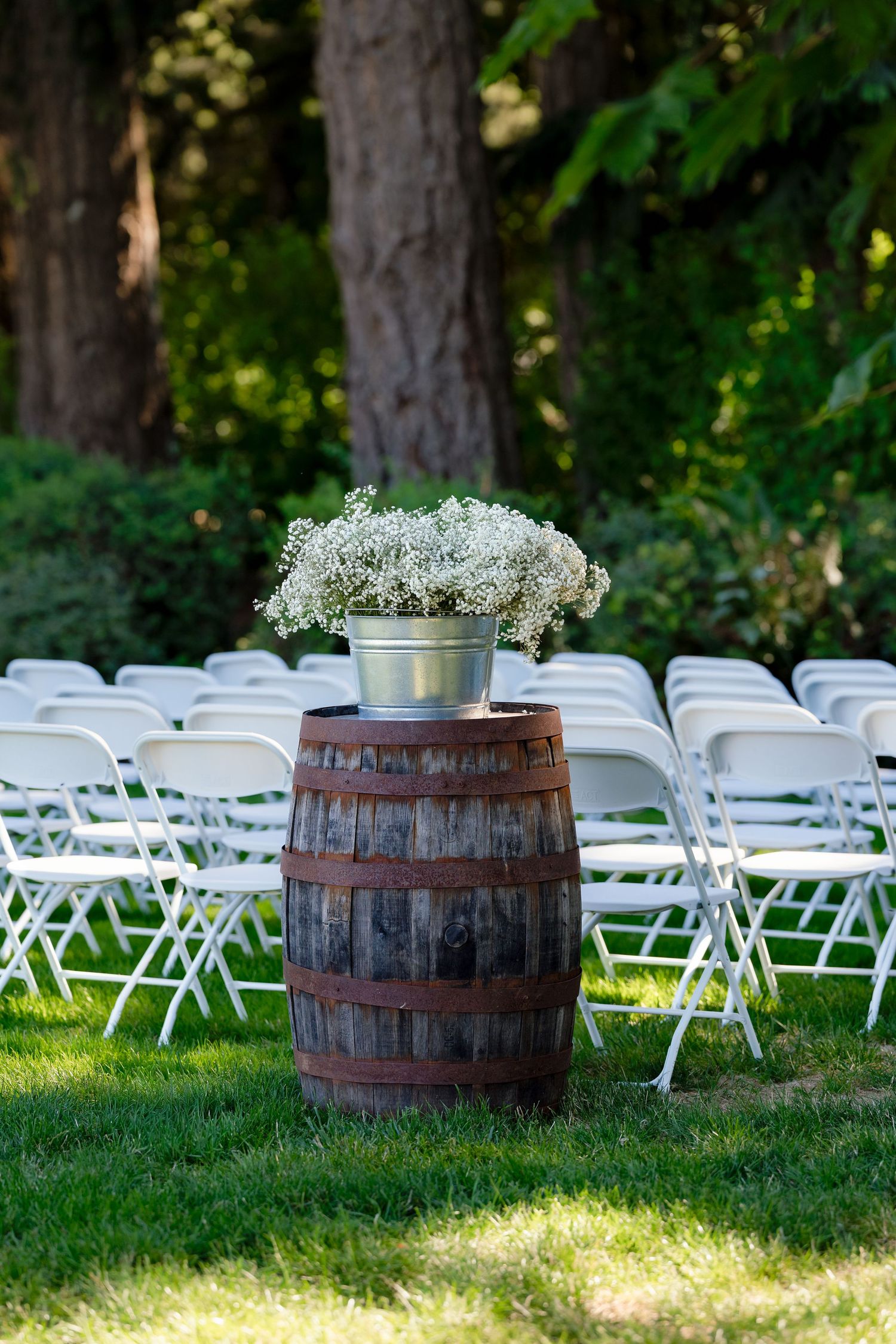 Rustic wooden barrel with white baby's breath flowers in silver bucket beside white chairs at outdoor wedding ceremony.