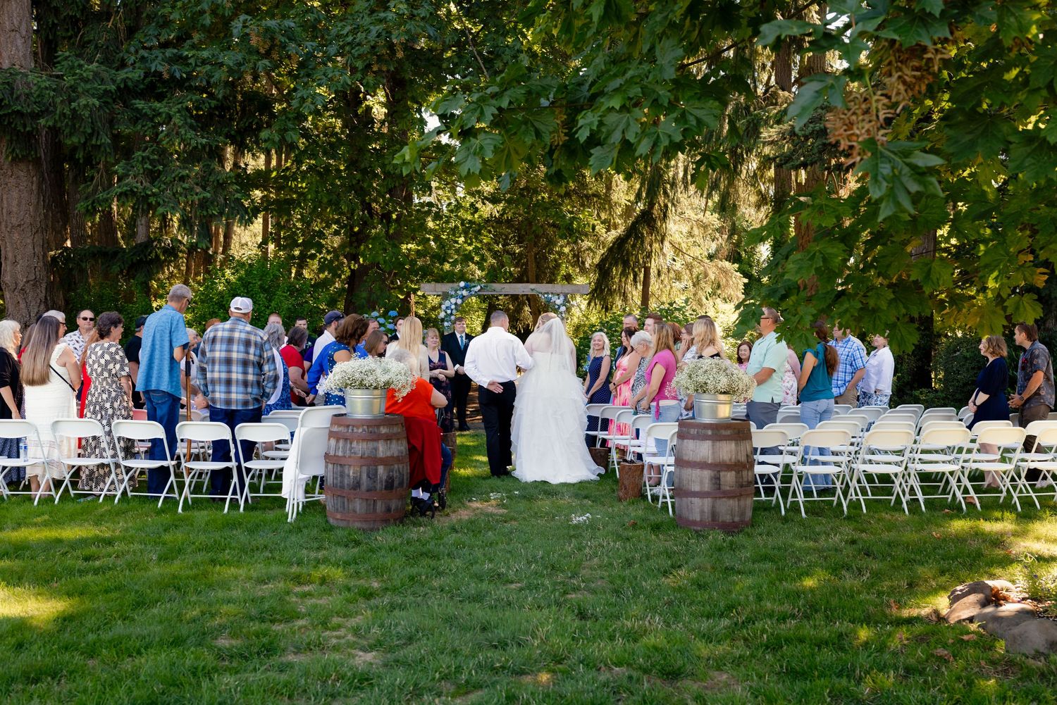Outdoor garden wedding ceremony with guests seated in white chairs flanking barrel aisle markers beneath tall trees.