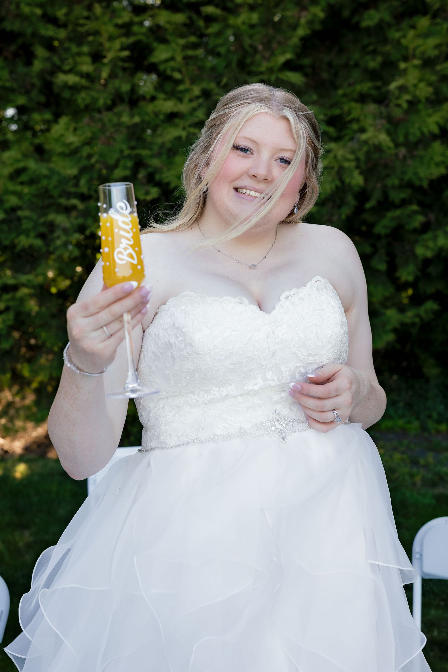Smiling bride in white strapless gown holding a champagne flute outdoors.