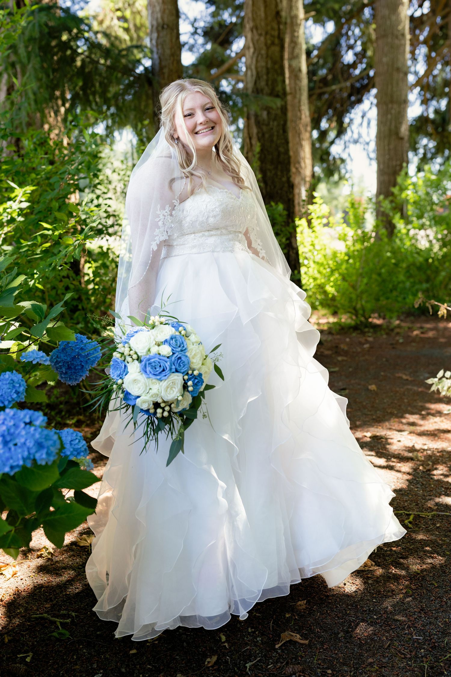 Smiling bride in a ruffled white wedding gown with veil holds blue and white bouquet among hydrangeas.