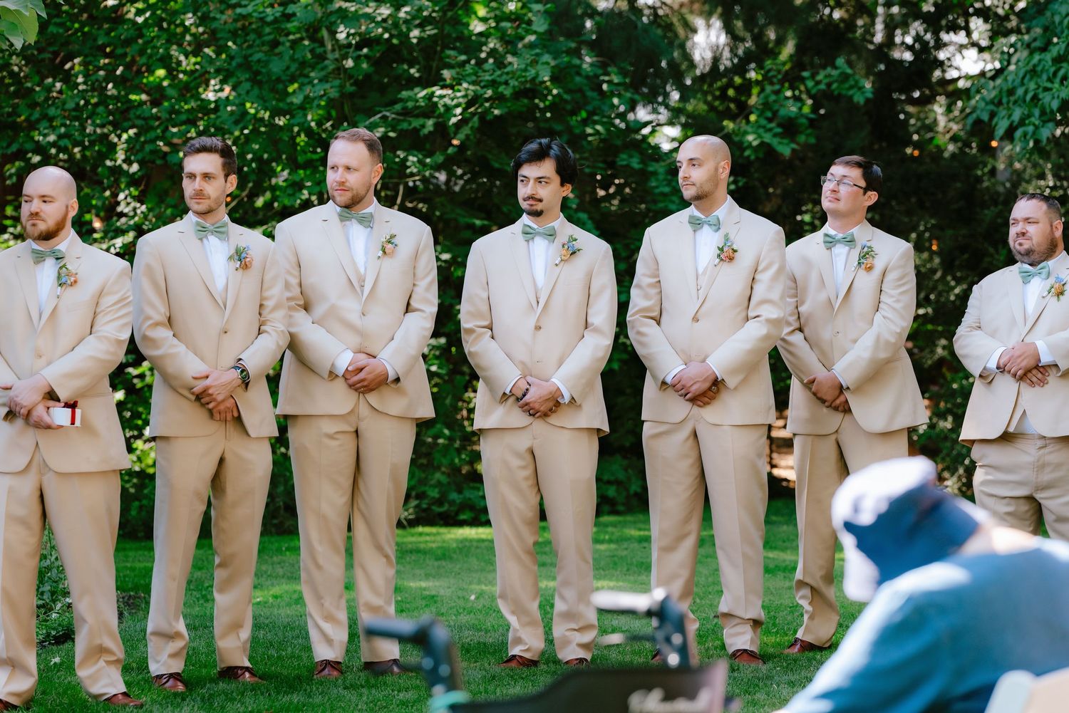 Groomsmen in matching tan suits with boutonnieres stand at outdoor garden wedding ceremony.