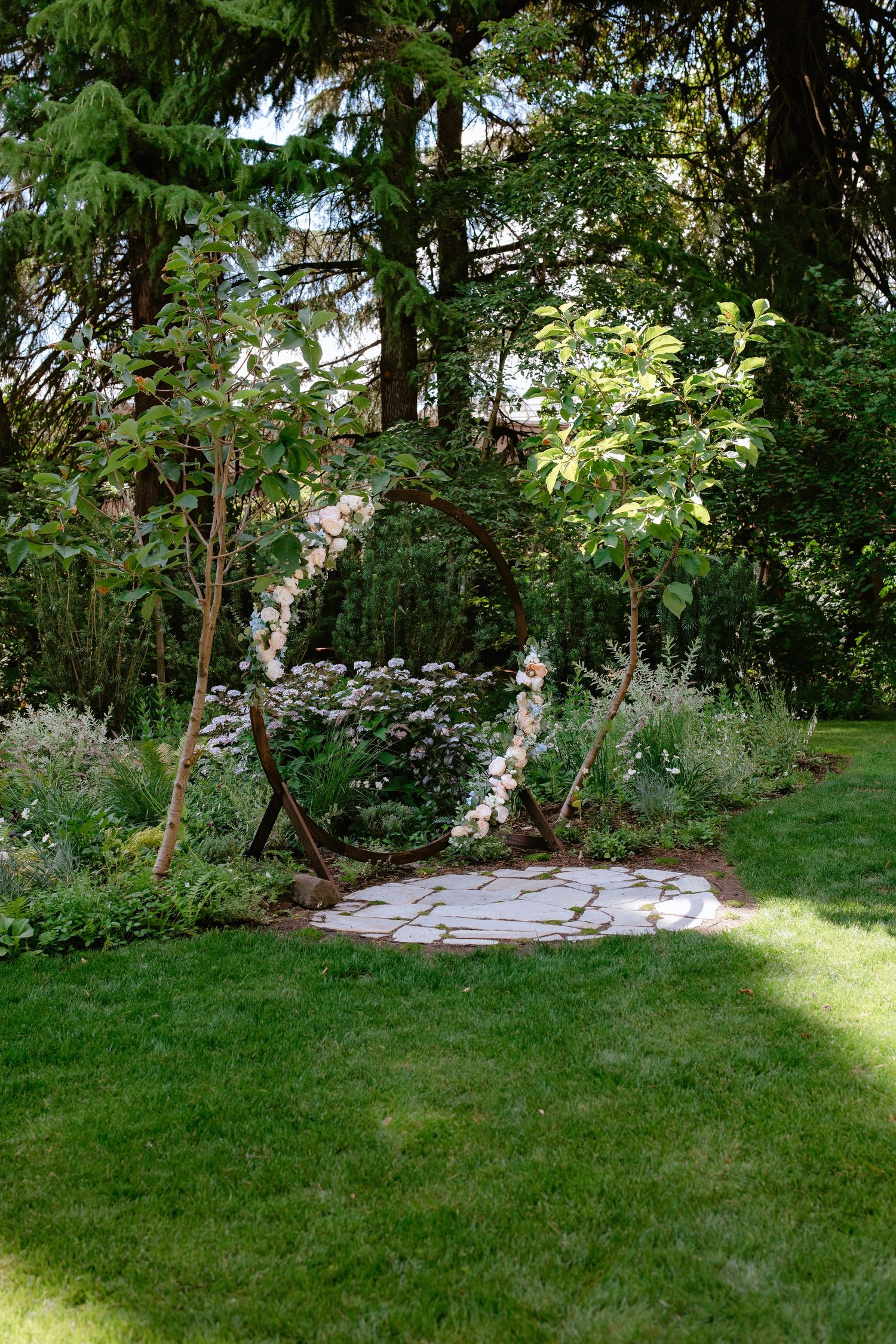 Circular wooden wedding arch adorned with pink flowers on stone patio in garden setting.