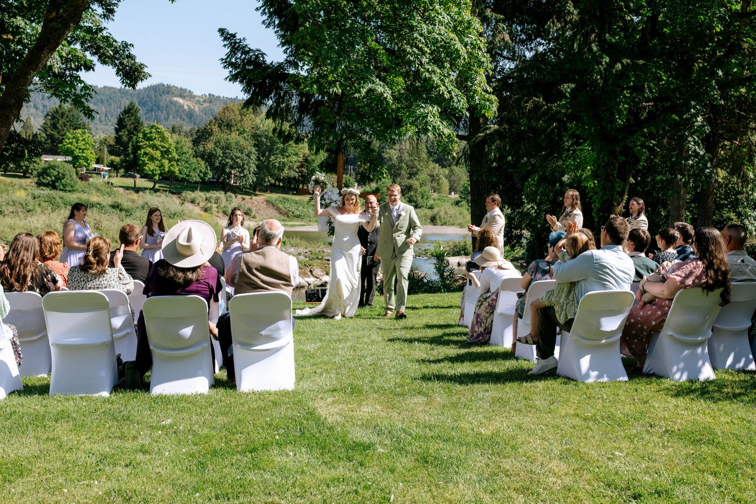 Outdoor wedding ceremony on a sunny lawn with guests seated in white chairs facing the couple near a scenic river.