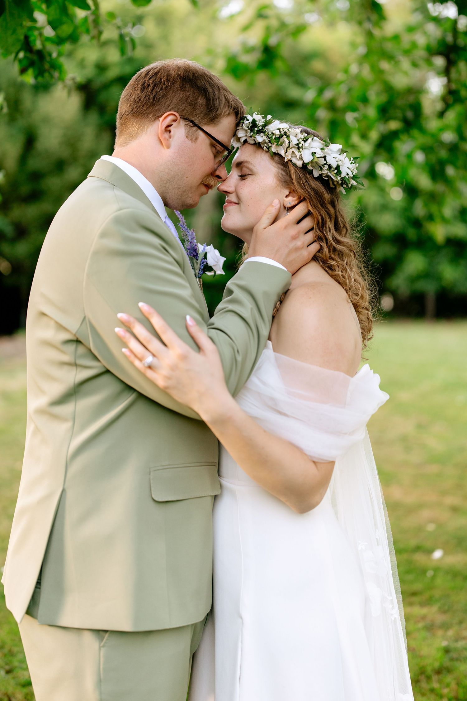 Groom in sage suit tenderly holding bride's face as they share an intimate moment in a garden.