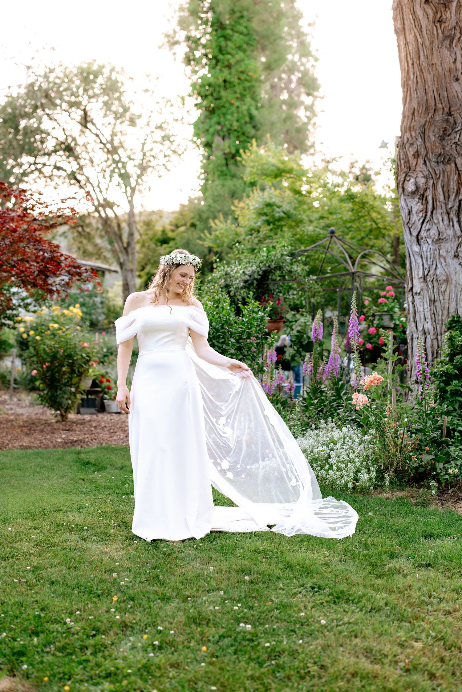 Bride in white off-shoulder gown with floral crown twirls veil in colorful garden at sunset.