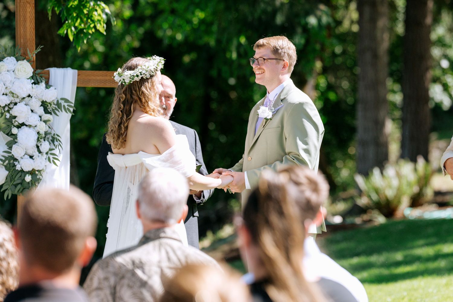 Bride in floral crown and groom in sage suit exchange vows at an outdoor garden wedding ceremony.
