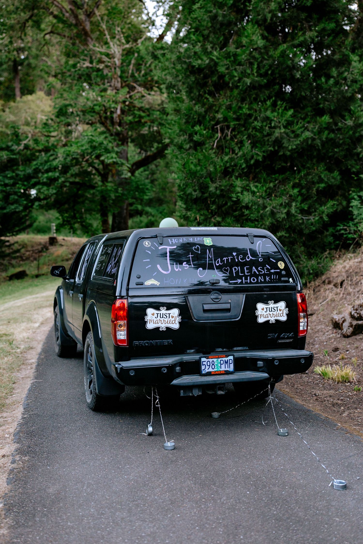 Black truck decorated with Just Married sign and tin cans driving away on a tree-lined road.