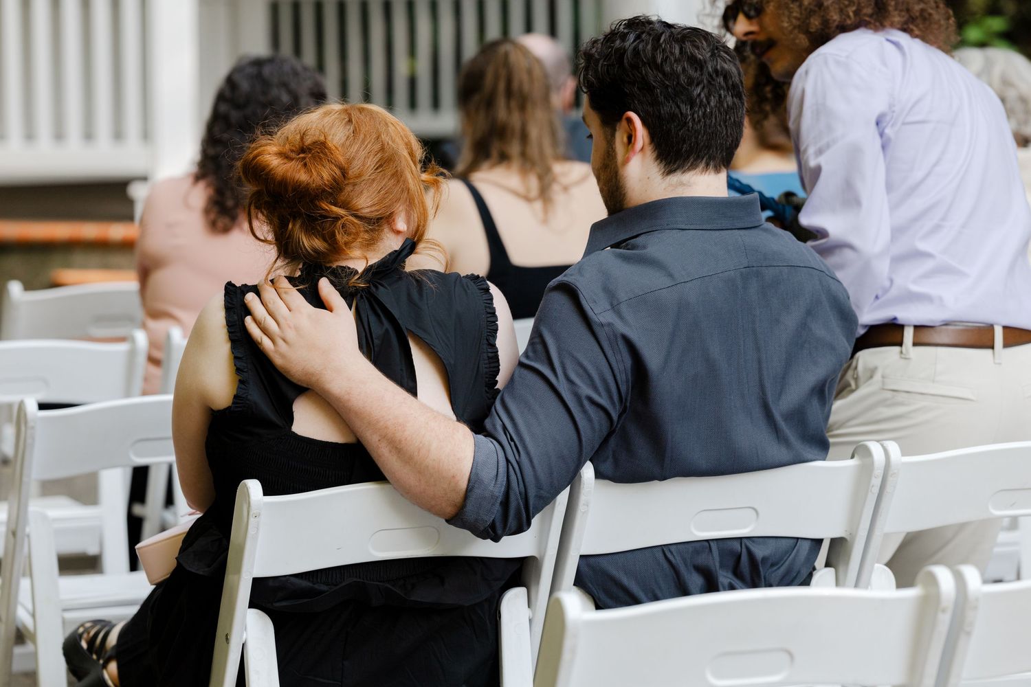 Couple embracing from behind at an outdoor wedding ceremony, seated on white chairs.