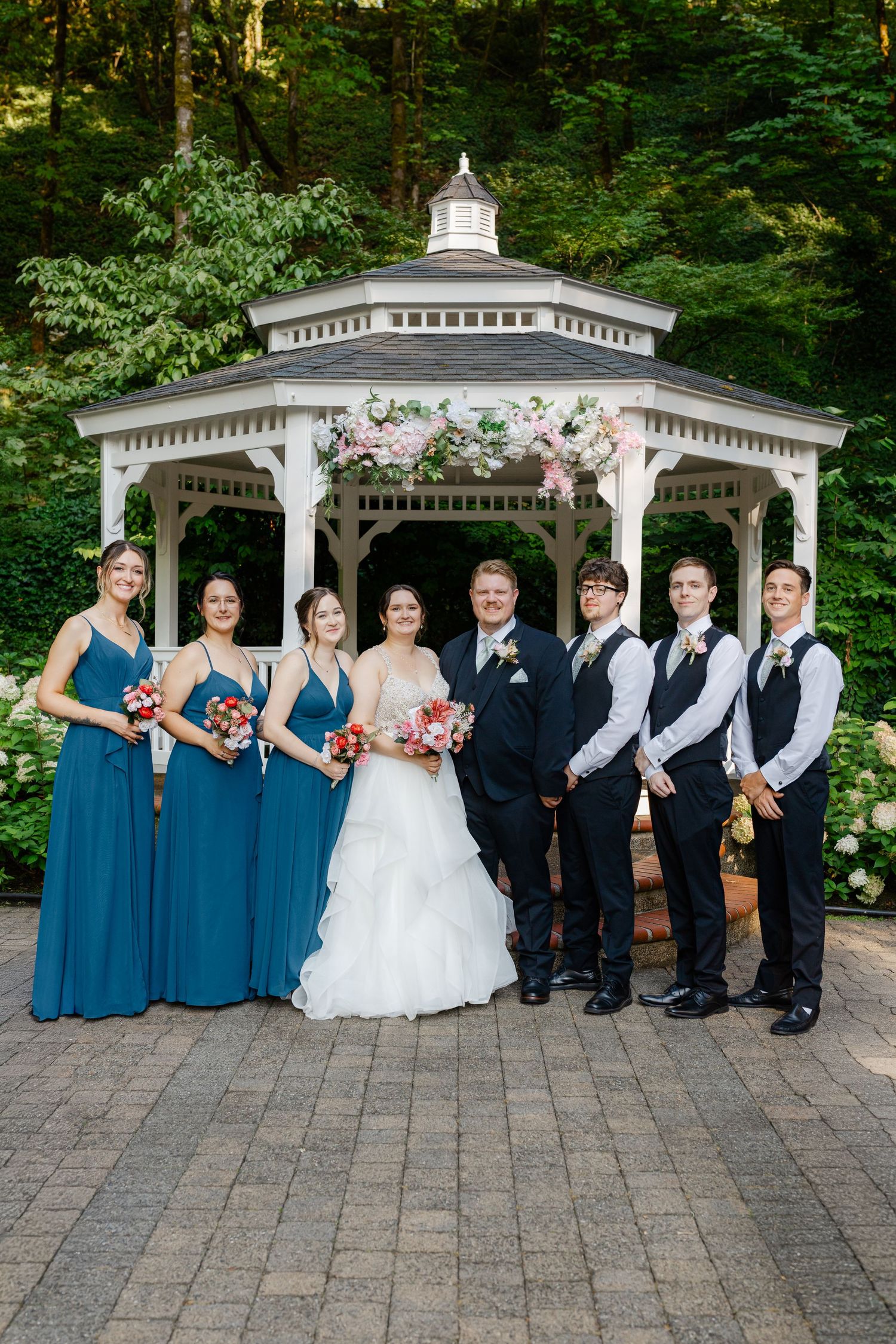 Wedding party in teal bridesmaid dresses and black suits posing before a flower-adorned gazebo.