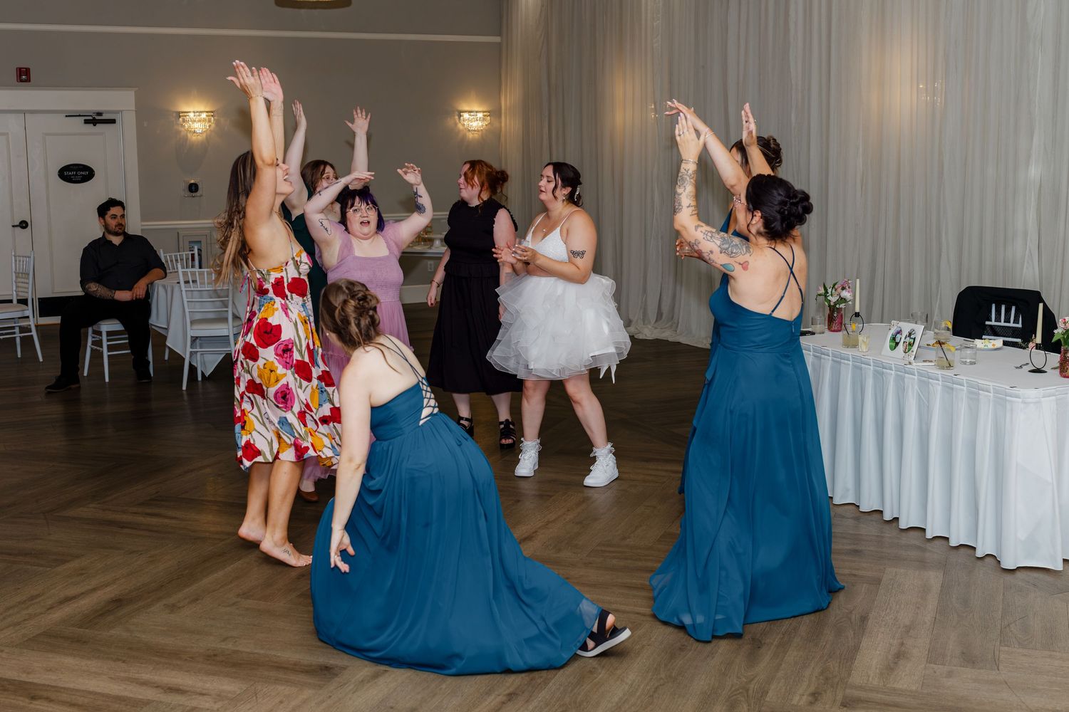 Bridesmaids in teal gowns dance energetically with the bride on a wooden reception dance floor.